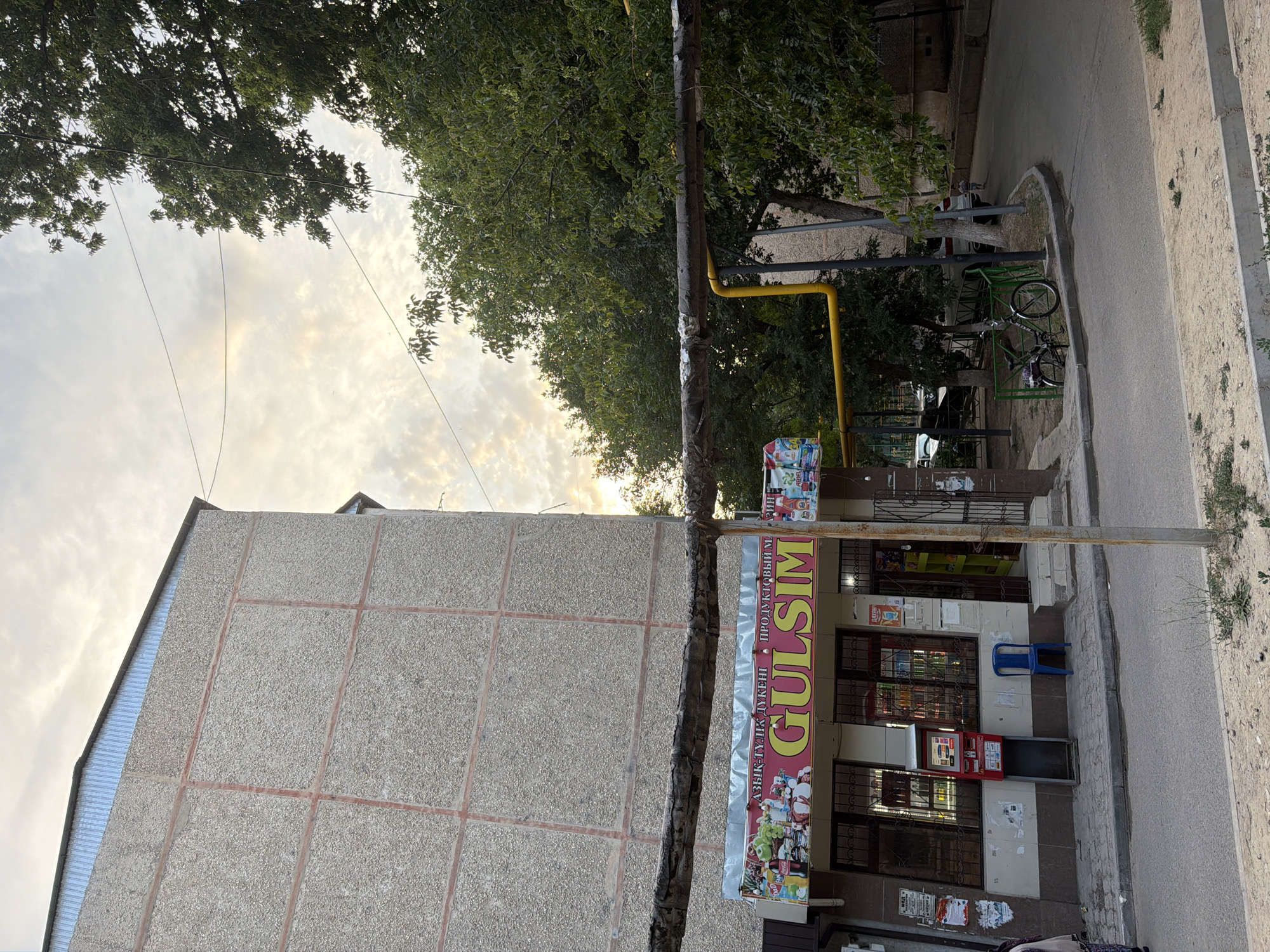 The image captures a street scene in Shymkent, Kazakhstan, likely during the daytime, as indicated by the bright sky. The sky is cloudy with some patches of light suggesting a potentially overcast or partly sunny day.
The main subjects in the foreground are a building with a textured, concrete facade and a storefront. The building on the left is large and imposing, with a metal-clad section on its left side. Adjacent to this building is a street and sidewalk. A utility pole stands prominently in the center of the image, with a yellow gas pipe running along it.
The storefront, labeled "GULSIM" in large, bold yellow lettering on a red banner, appears to be a small shop or market, possibly a convenience store given the products visible through the windows. The windows are covered with security bars. In front of the store, a blue plastic chair sits near the entrance. There's also a red and silver machine, possibly an ATM or a payment terminal, located outside the store. The storefront is adorned with various colorful signs and advertisements, some of which are partially visible.
In the background, trees with green foliage suggest a residential or urban area with some greenery. A bicycle is parked near a green metal fence. Further in the background, another building is visible. The overall scene depicts a typical street view in a city, with a focus on the storefront and the surrounding environment. The image is oriented vertically, meaning the scene is viewed from a slightly elevated or tilted perspective. No people are clearly visible in the image, but the presence of a store and a bicycle implies a populated area.