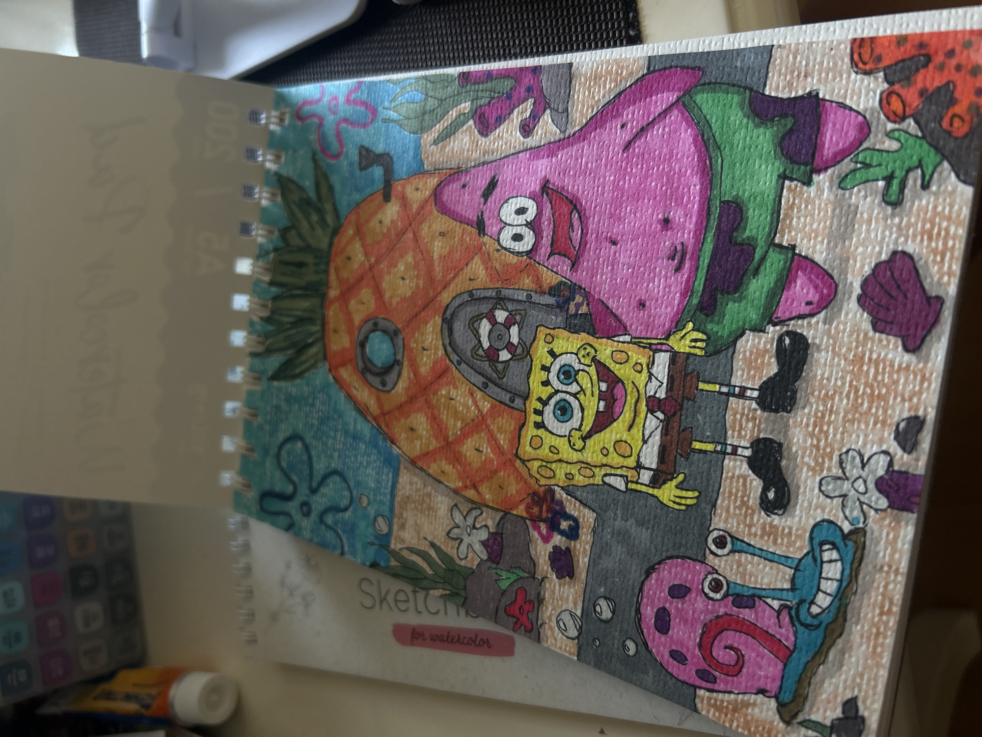 The image displays a close-up, overhead view of a drawing on a spiral-bound sketchbook page. The drawing features characters from the animated series SpongeBob SquarePants, specifically SpongeBob, Patrick, and Gary. They are depicted in their underwater environment with SpongeBob's pineapple house in the background. The drawing is rendered in colored pencils, with vibrant colors and visible pencil strokes. The sketchbook page is placed on a textured surface, possibly a table, with other objects in the background, including art supplies. The text "Sketch" and "for watercolor" is visible on the left side of the sketchbook page. The overall impression is of a child's drawing or a hobbyist's artwork. There is no information in the image that suggests a location in Khyrdalan, Azerbaijan.