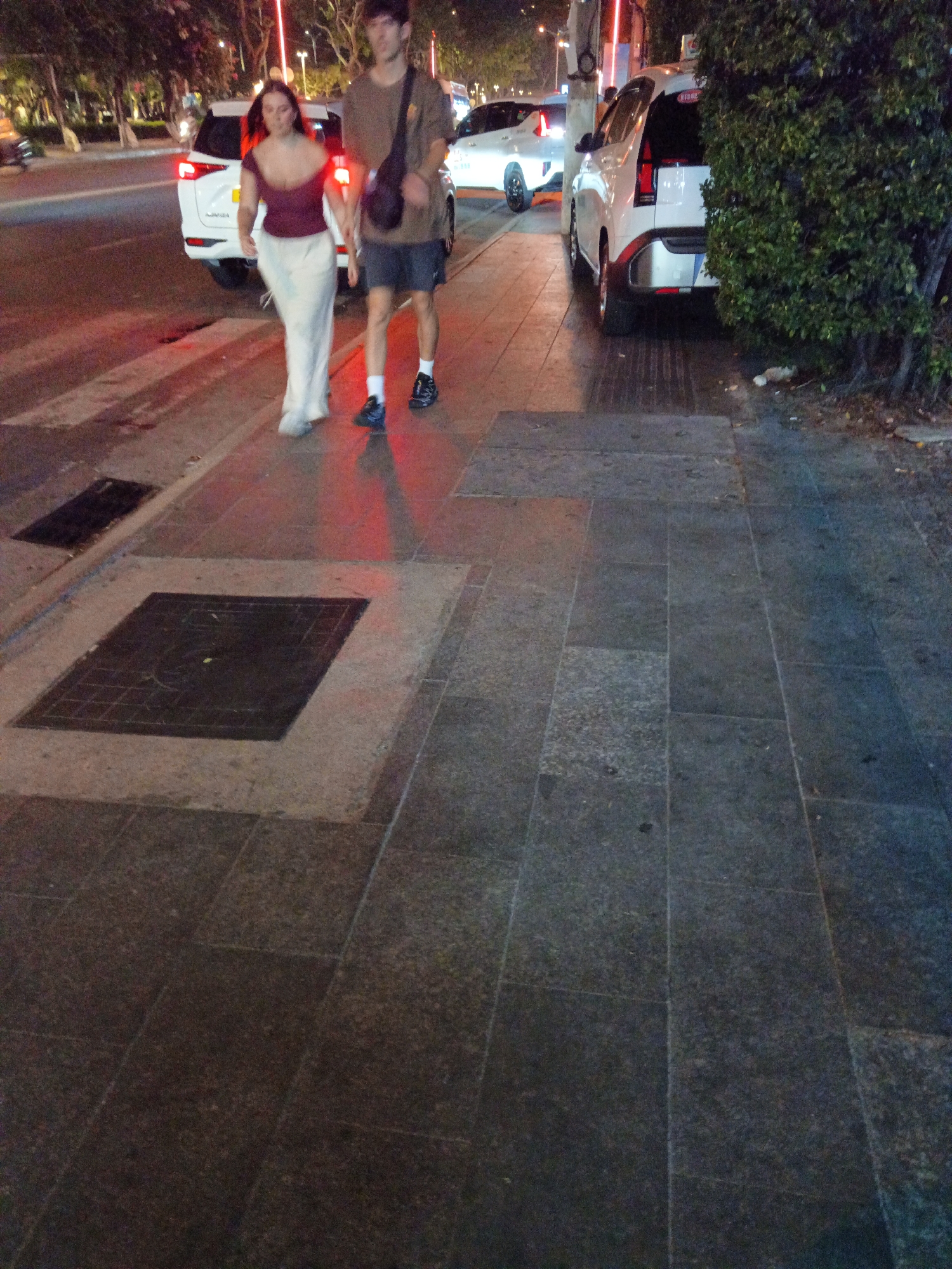 In Nha Trang, Vietnam, at night, two individuals, a woman and a man, walk on a sidewalk adjacent to a street. The woman, wearing a burgundy off-the-shoulder top and a white flowing skirt, walks slightly ahead of the man. The man, dressed in a brown t-shirt and dark shorts, carries a dark bag over his shoulder. They are walking away from the viewer, towards the right of the frame.
Behind them, a white SUV is parked on the street, its headlights and taillights illuminated. Across the street, another white car is visible. The sidewalk is paved with large, dark grey stone tiles. To the right of the sidewalk, a dense green bush is prominent. In the foreground, a square metal grate is set into the pavement, and to its left, a rectangular metal drain cover is also visible. The overall lighting suggests an urban environment at night, with artificial lights casting shadows and reflections.
