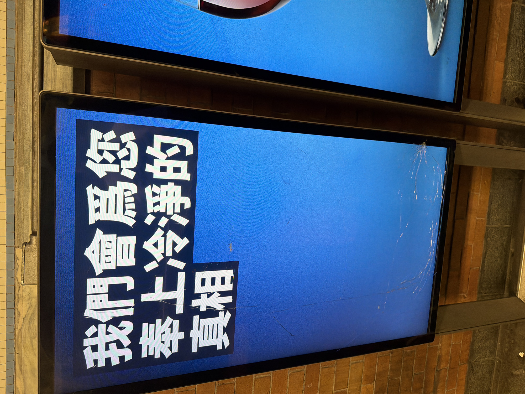 The image shows two digital display panels mounted side-by-side on a wall with brickwork visible in the background. The lower panel is cracked in the upper right corner. The screen displays text in white, vertically aligned in a blue background. The text is in Chinese. The upper panel is also blue and has some sort of reflective silver object. The location appears to be an indoor setting, possibly a subway station in New York City.