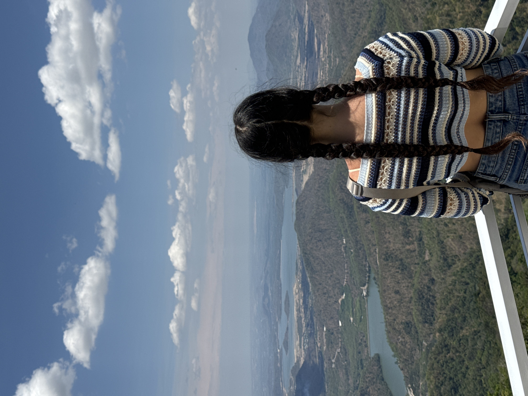 A woman with dark hair in braids is standing on a high vantage point, overlooking a landscape in Nam Hải, Vietnam. She wears a striped blue and white top and denim jeans. The view includes a series of lakes or reservoirs nestled between forested hills. The sky is blue with scattered white clouds. The woman is standing near a white railing.