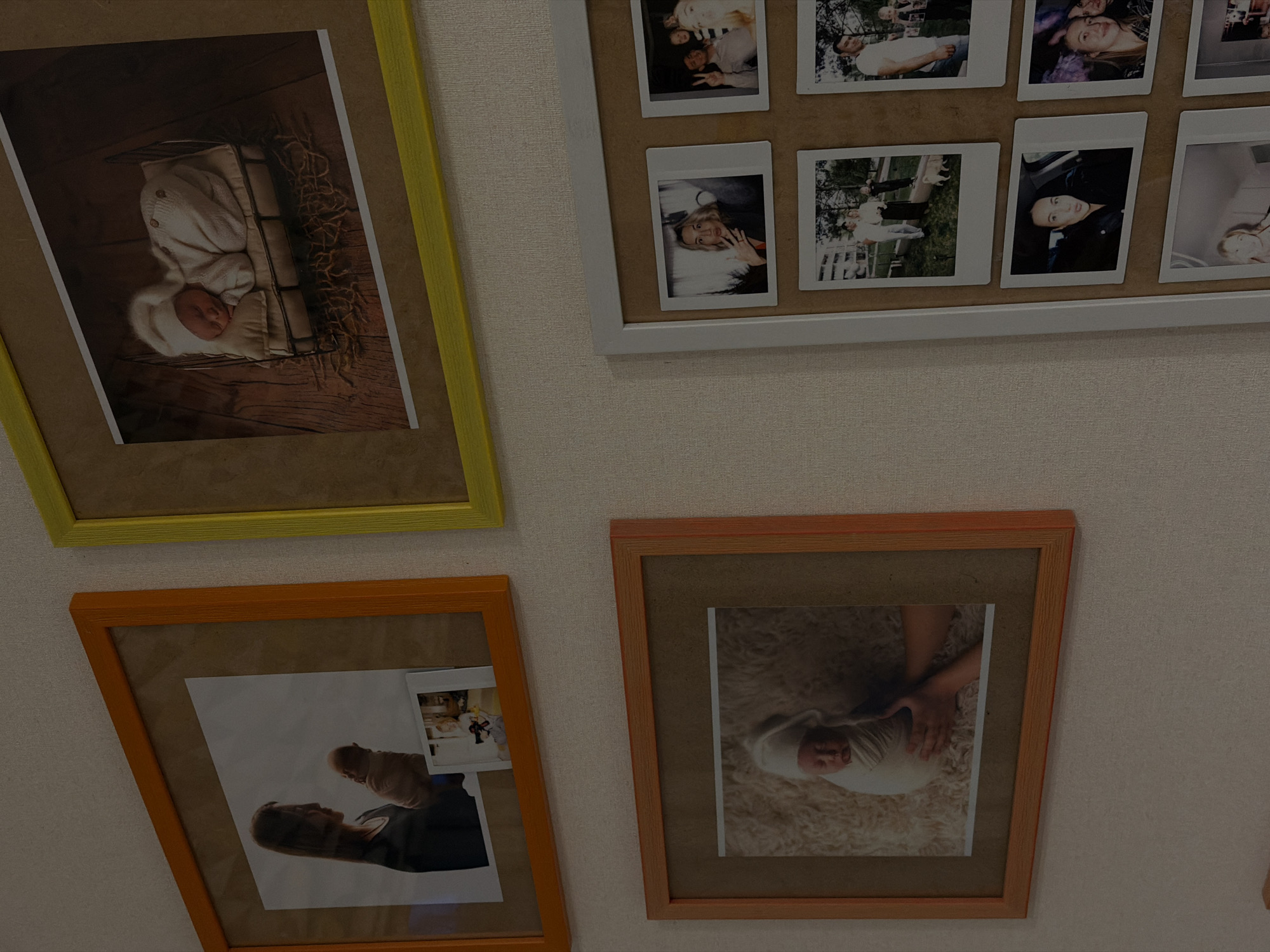The image captures a wall display of framed photographs in a home setting in Sofiivska Borshchahivka, Ukraine. Four main frames, varying in size and color, are mounted on a light-colored, textured wall, showcasing a collection of personal memories.
In the upper left, a vibrant yellow frame encloses a portrait of a newborn baby. The baby is depicted sleeping peacefully, swaddled in a white knitted outfit and a matching hat, lying within a rustic basket set against a dark, wooden background.
To the upper right, a large white frame contains multiple smaller, polaroid-style photographs. These images feature various individuals and groups, including adults smiling in group shots, a woman making a playful expression with her hand near her face, and other candid moments, some appearing to be outdoor scenes and others indoors.
The lower left features an orange frame holding a larger print and a smaller, overlaid polaroid. The main print shows a woman with long hair looking upwards while gently holding a baby against her chest, both silhouetted against what appears to be a bright sky. A smaller, partially visible polaroid hints at an indoor celebratory event.
In the lower right, a second orange frame displays a close-up photograph of a baby lying on a soft, fluffy white blanket. An adult's hand is tenderly placed on the baby, who is dressed in light-colored clothing and appears to be asleep or resting.
The collection of photos predominantly features themes of family, newborns, and cherished personal moments, suggesting a display of significant life events and relationships. No discernible text is visible on any of the frames or photographs.