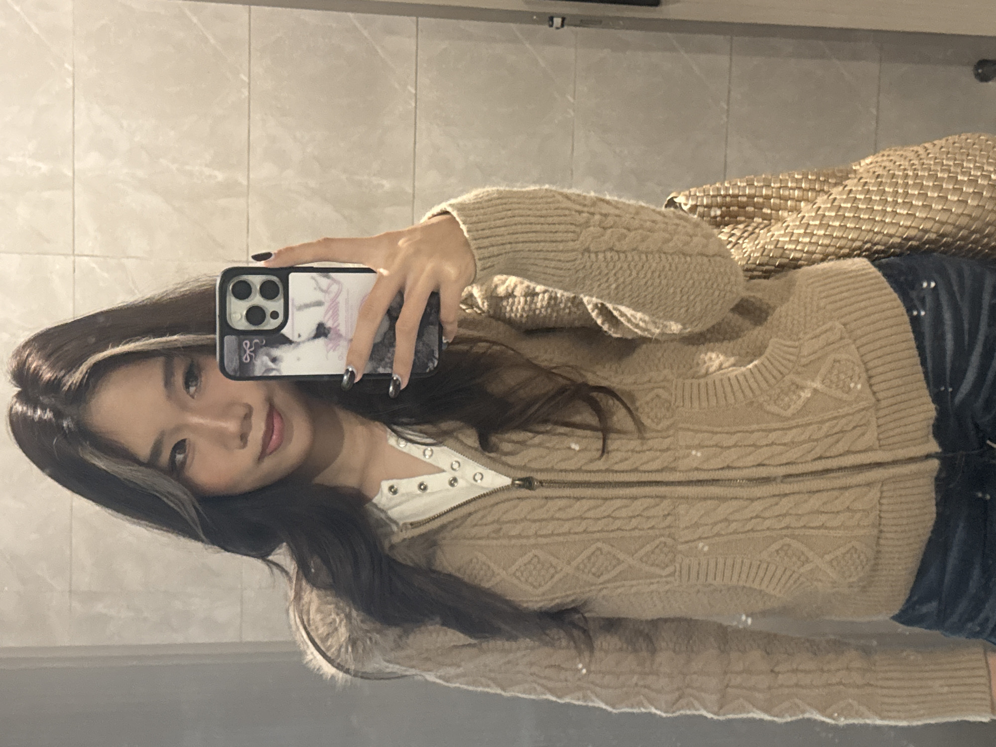 An East Asian female is depicted from the waist up in a mirror selfie taken indoors. She has long, dark brown hair with a lighter streak on the left side, and a subtle smile. Her right hand holds a dark-cased smartphone, featuring a multi-lens camera module and a case design incorporating a black-and-white image and a bow icon. Her fingernails are painted black. She wears a beige cable-knit hooded cardigan with a fur-trimmed hood over a white Henley-style shirt with four visible buttons. Dark blue denim jeans are visible below the cardigan, along with what appears to be a light brown woven bag slung over her right shoulder. The background consists of light-colored, possibly beige or off-white, tiled walls with horizontal grout lines. There are no discernible visual cues within the image to specifically identify the location as Nan'an District, China.
