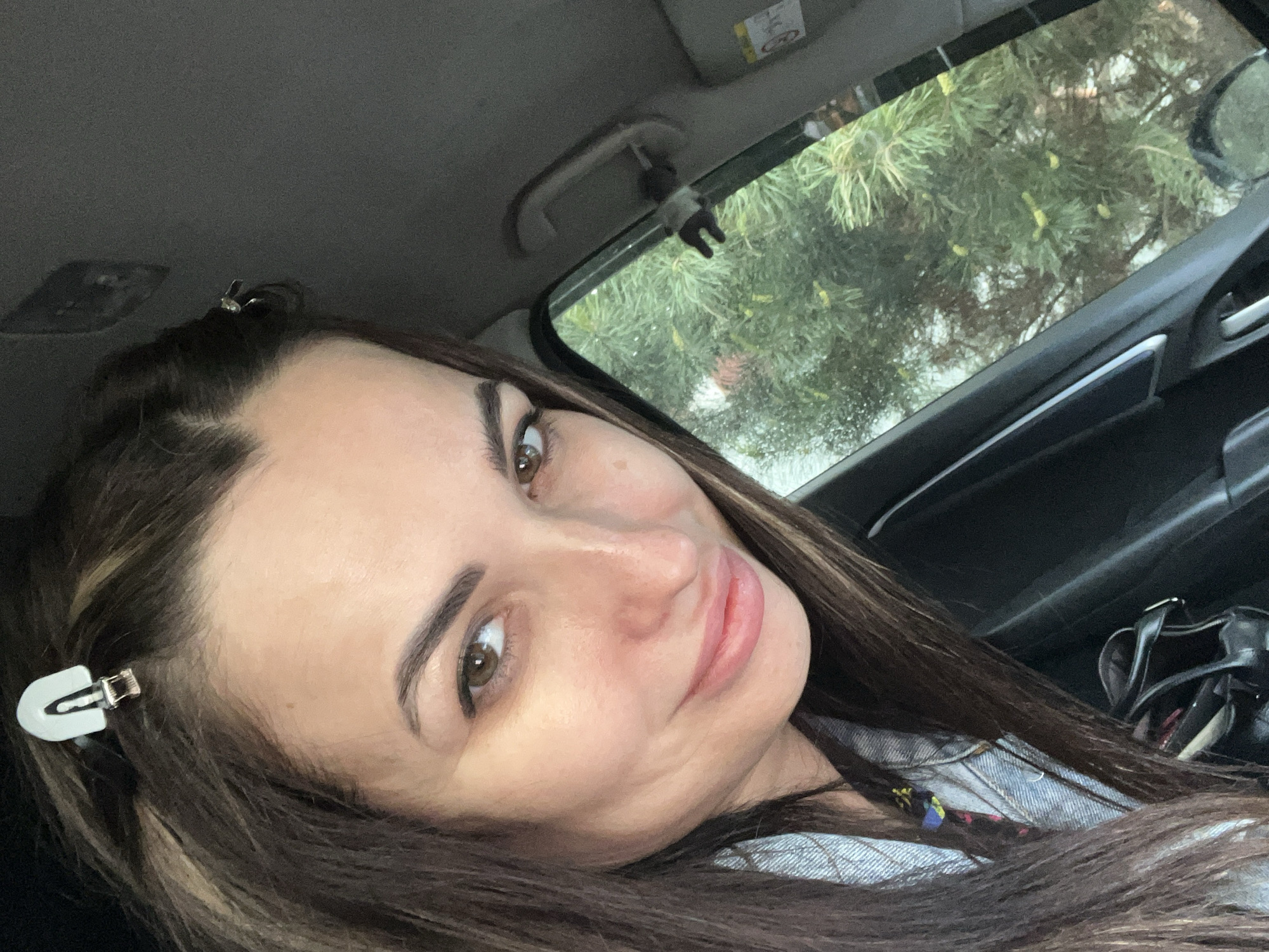 A selfie shows a woman with brown hair and fair skin, wearing a blue denim jacket, inside a car. She has two white hair clips in her hair, one above each eye. The view through the car window shows green pine trees. The interior of the car is visible, including a black handbag, the window frame, and the car's ceiling. The location is Unknown, Unknown.