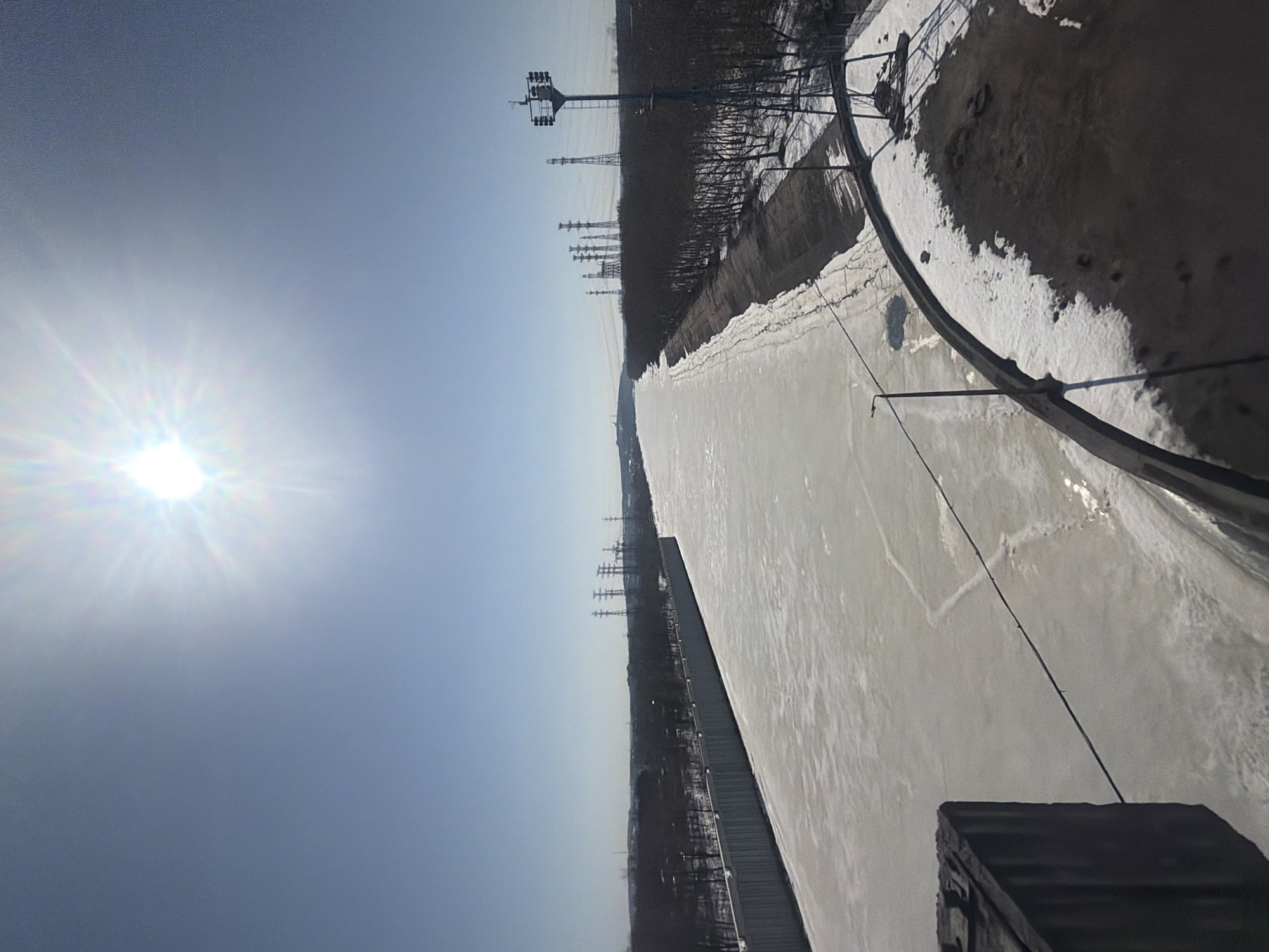 The image depicts a frozen reservoir with power lines in Chaikovsky, Russia. The sun is visible in the sky. A fence is present on the right side of the frame.