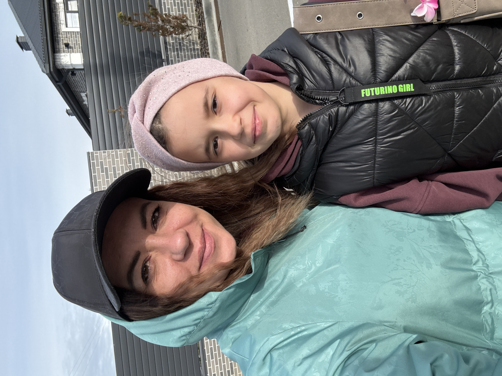 The photograph captures a woman and a young girl posing together. The woman is wearing a black baseball cap and a light blue hooded jacket. The girl is dressed in a black puffer jacket with the words "FUTURINO GIRL" on it, and a light pink headband. The background reveals a building with a gray exterior and the presence of a paved surface, suggesting an urban environment. The location of the photo appears to be Taman, Russia.