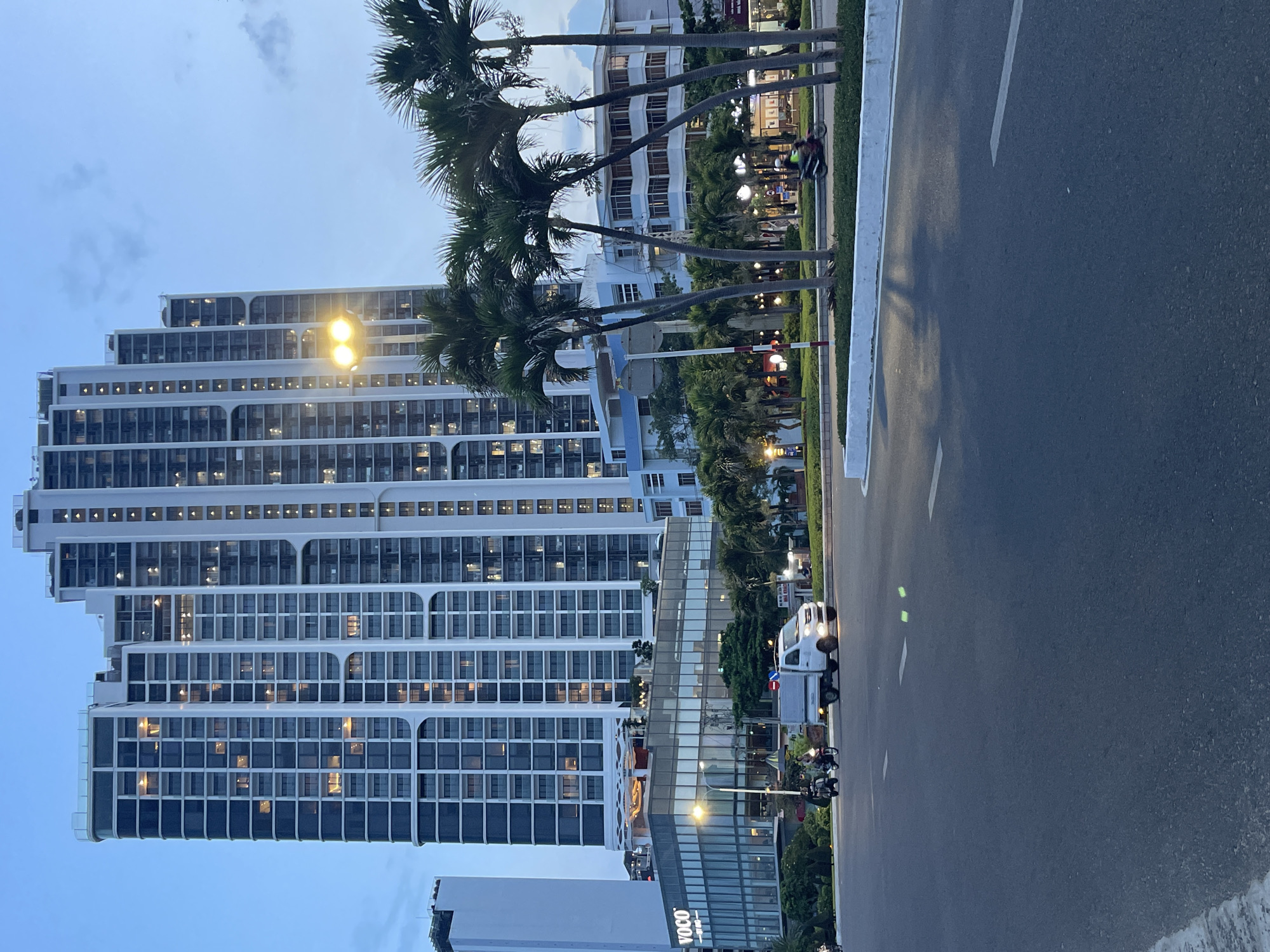 This is a photograph of the waterfront area in Nha Trang, Vietnam, likely at dusk or early evening. The scene is dominated by a tall, modern, white high-rise building with many windows lit from within. In front of this building, there is a lower, glass-fronted building with the "VOCO" logo visible on its facade. Palm trees line the foreground, and a paved road with white lane markings occupies the bottom half of the image. Several motorcycles and a small white truck are visible on the road. The sky is a clear, pale blue.