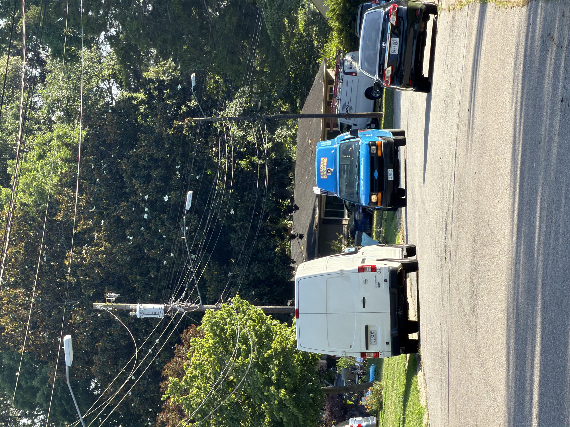 The image captures a residential street scene in Virginia Beach, Virginia, USA, on a clear, sunny day. In the foreground, the asphalt road is visible with tire tracks and shadows cast by overhead power lines and trees. On the right side of the road, a black Toyota Corolla is parked, its license plate reading "DWEY BCH". Further up the street, a bright blue Chevrolet van, identified as an ice cream truck with "ICE CREAM" text and "CHILDREN CROSSING" signage, is driving towards the camera. To the left of the ice cream truck, a white Nissan NV cargo van is also parked.
The background features a typical suburban residential setting with houses and mature trees lining the street. Overhead, a complex network of power lines and utility poles crisscrosses the sky, with streetlights visible. The trees are lush and green, indicating a warm season. The overall impression is a quiet, ordinary street in a residential neighborhood during daylight hours. No people are clearly visible in the image.