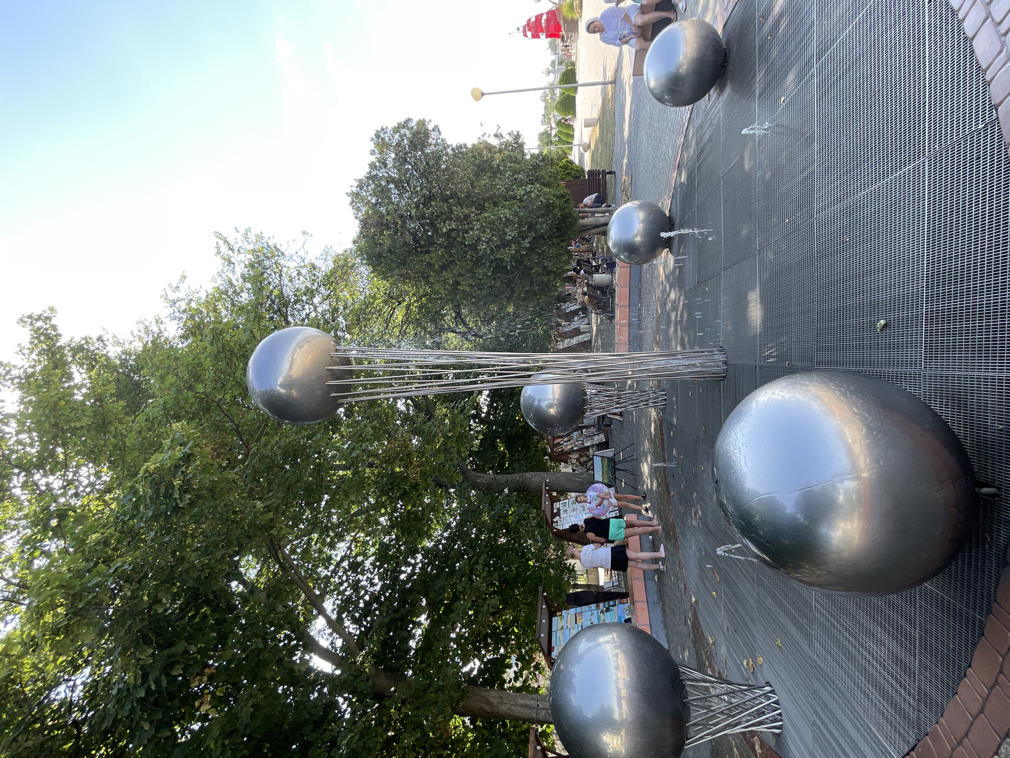 The image shows a public space with a water feature. The water feature consists of large, metallic spheres with thin rods extending upwards from them, from which water sprays. Several people are visible, including a group of young people standing near the water feature and a man sitting on a bench in the background. The scene is outdoors, with lush green trees providing shade. The ground is paved with a metal grate in the area of the water feature, surrounded by brickwork. In the background, there are more trees, a lamppost, and a red structure that resembles a sail, possibly part of a decorative element. The time of day appears to be daytime, with bright sunlight casting shadows. The weather seems pleasant, conducive to outdoor activities. The location is identified as Anapa, Russia. The overall mood conveyed is one of leisure and enjoyment of a public amenity on a sunny day. There is no visible text in the image.