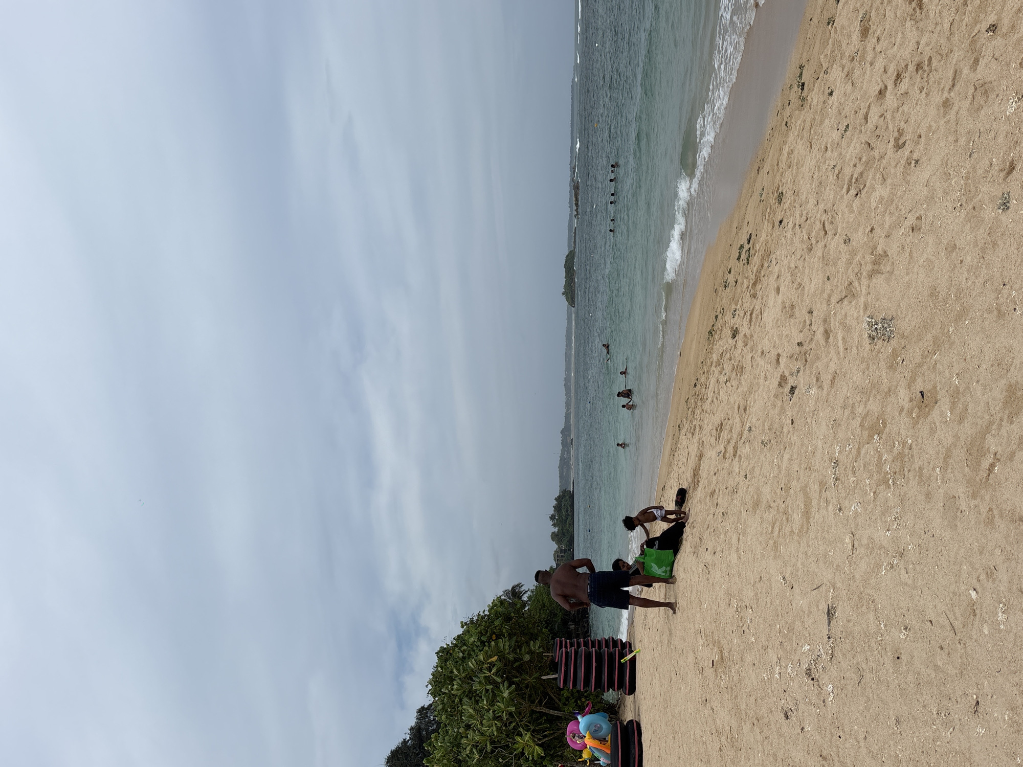The image depicts a beach scene in Weliweriya, Sri Lanka. A group of people, including adults and a child, are gathered on the sand near the water's edge. A man is standing near some personal belongings, including what appears to be inflatable tubes and a green bag. In the background, several people are in the water. The sky is overcast. The sand is light-colored, and the water is a greenish-blue. Vegetation is visible in the lower left of the image.