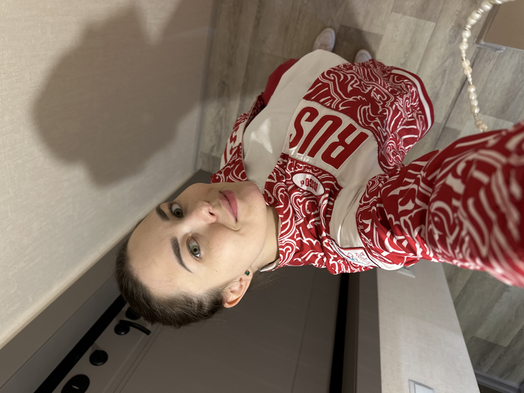 A young woman with light skin, dark hair pulled back, and striking green eyes looks directly at the camera with a subtle smile in a selfie-style photograph. She is wearing a distinctive red and white jacket or tracksuit, featuring a swirling pattern and large white letters spelling "RUS" (likely an abbreviation for Russia) on the chest area. A small label on the sleeve, partially visible, appears to be from a sports outfitter, possibly ZASPORT, and includes a small emblem. She also wears a small green stud earring.
The setting appears to be an indoor hallway or room with light-colored, possibly textured, walls and a light-toned wooden or wood-effect tiled floor. A dark-colored door with black handles and locks is visible to her left. Her white sneakers are visible on the floor, and a decorative pearl-like beaded strap, possibly from a bag or accessory, hangs on the right side of the frame. The lighting suggests an indoor environment without specific time-of-day cues. The image is taken from a slightly high angle, typical of a mirror or handheld selfie. Given the context of Perm, Russia, the attire suggests a connection to national sports or representation.