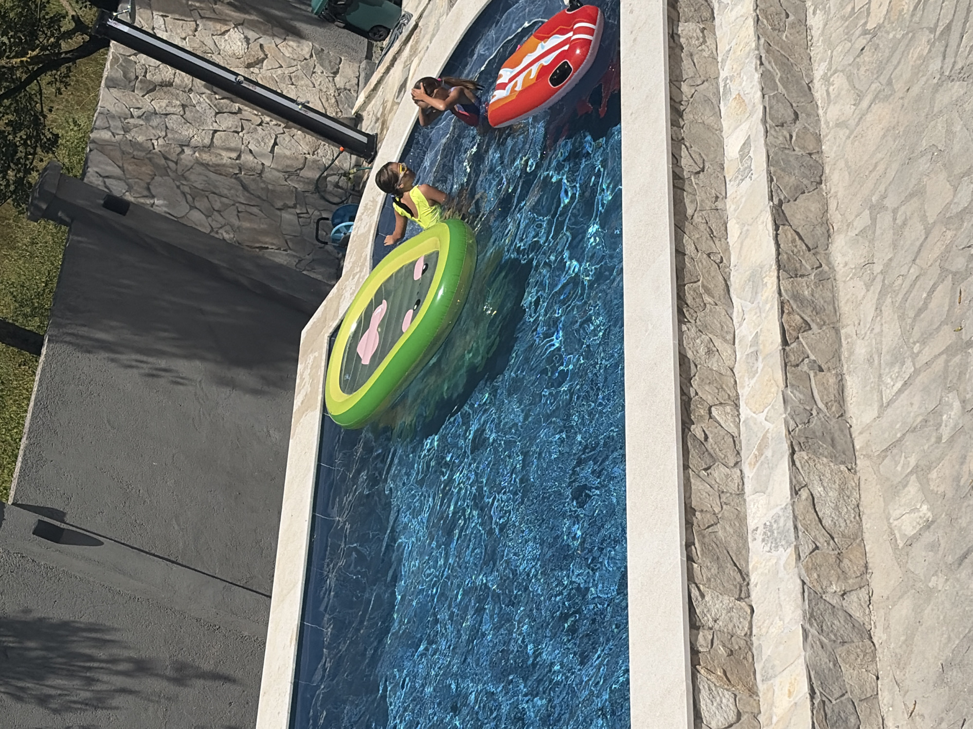 Two young girls are in a swimming pool. One girl, wearing a yellow swimsuit and goggles, is standing near a green inflatable raft shaped like a sea creature. The other girl, wearing a dark swimsuit, is in the water near a red and orange inflatable. The pool is surrounded by stone steps and has a dark, textured wall in the background. The scene appears to be a private residence in Lastva Grbaljska, Montenegro.