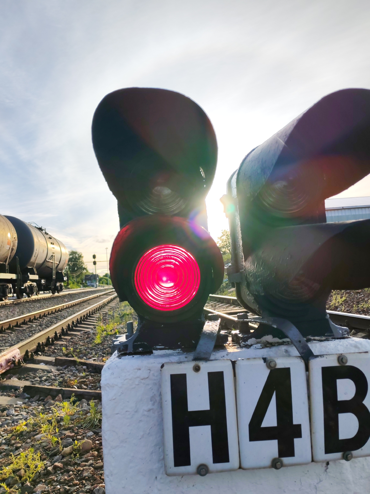 A close-up shot of a train signal in Bol'shie Mkhi, Russia. The signal displays a lit red light. In the background, a train car is visible on a train track. The signal is labeled "H4B." The sky is bright with the sun partly visible. The location appears to be alongside a railway. There are some plants between the rocks.