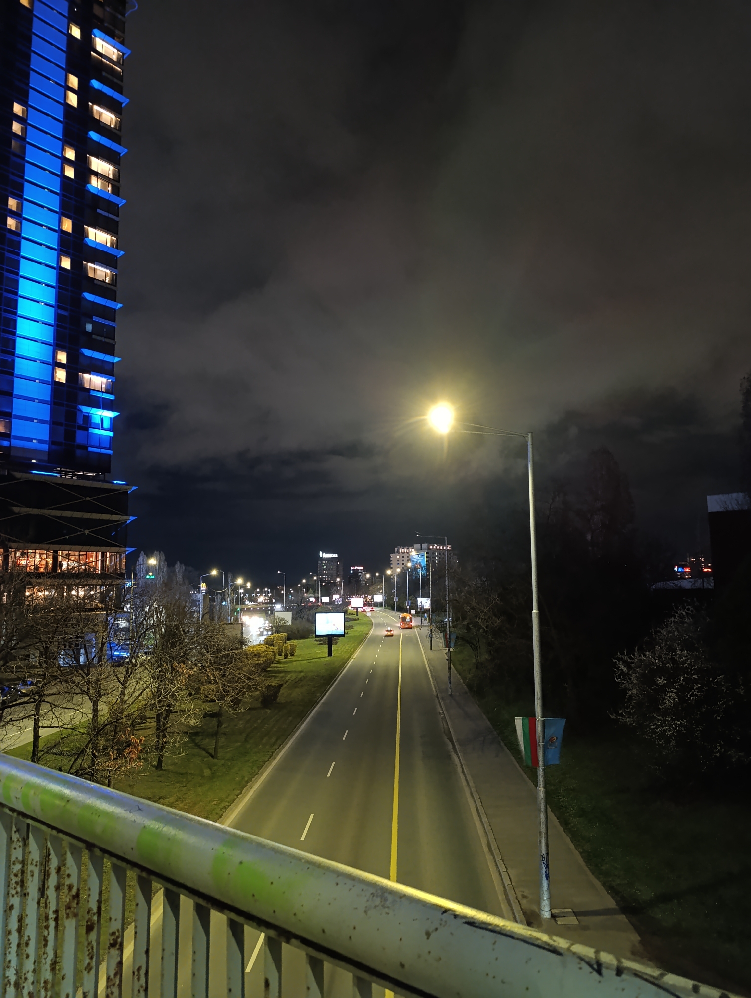 Here is a description of the image:
The image shows a nighttime view of a multilane road in Sofia, Bulgaria. A tall building illuminated with blue lights stands prominently on the left side of the frame, while the street stretches out into the distance, lit by streetlights. The road is divided by a yellow line, with cars visible in the distance. The sky is dark and cloudy. In the foreground, a metal railing suggests the image was taken from a pedestrian bridge. On the right side of the road, a flag with the colors green, white, and red, as well as an emblem, is attached to a light pole. There is greenery on either side of the road.