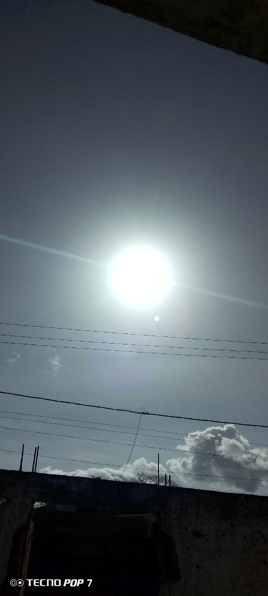 The image captures a bright, hazy sky with a prominent sun, possibly taken in Altagracia, Venezuela. Below the sun, several layers of utility lines stretch across the frame. In the lower portion, there's a glimpse of what appears to be the top of a building or structure with clouds visible behind it. The lower part of the structure appears dark and shadowed. The words "Tecno Pop 7" are visible in the bottom left corner of the image.