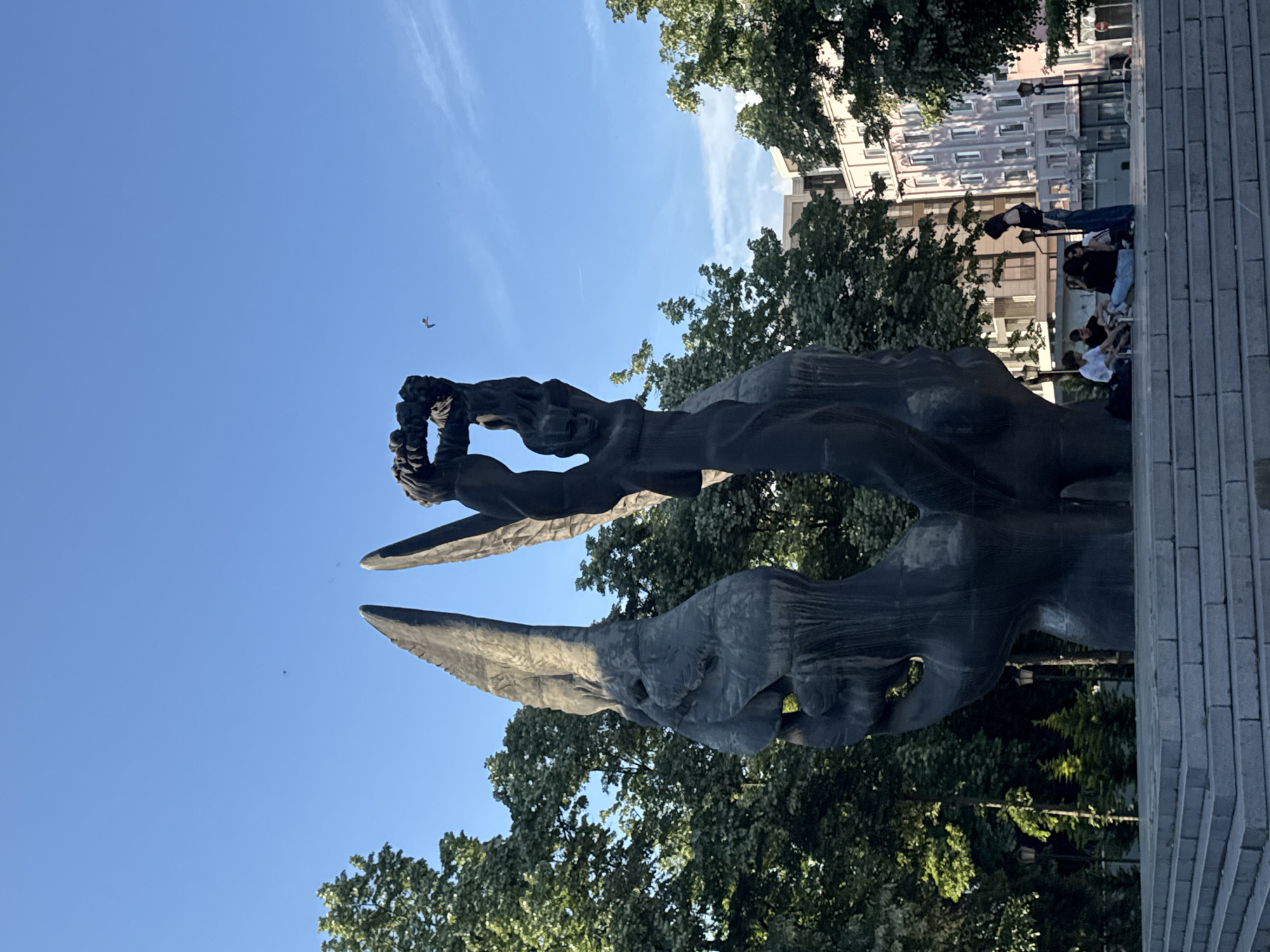 The image shows a dark-colored, abstract sculpture in Plovdiv, Bulgaria. The sculpture appears to feature human figures, with one figure holding a wreath above its head. The sky is blue, and trees are visible behind and around the sculpture. People are sitting on nearby steps. A building is partially visible in the background.