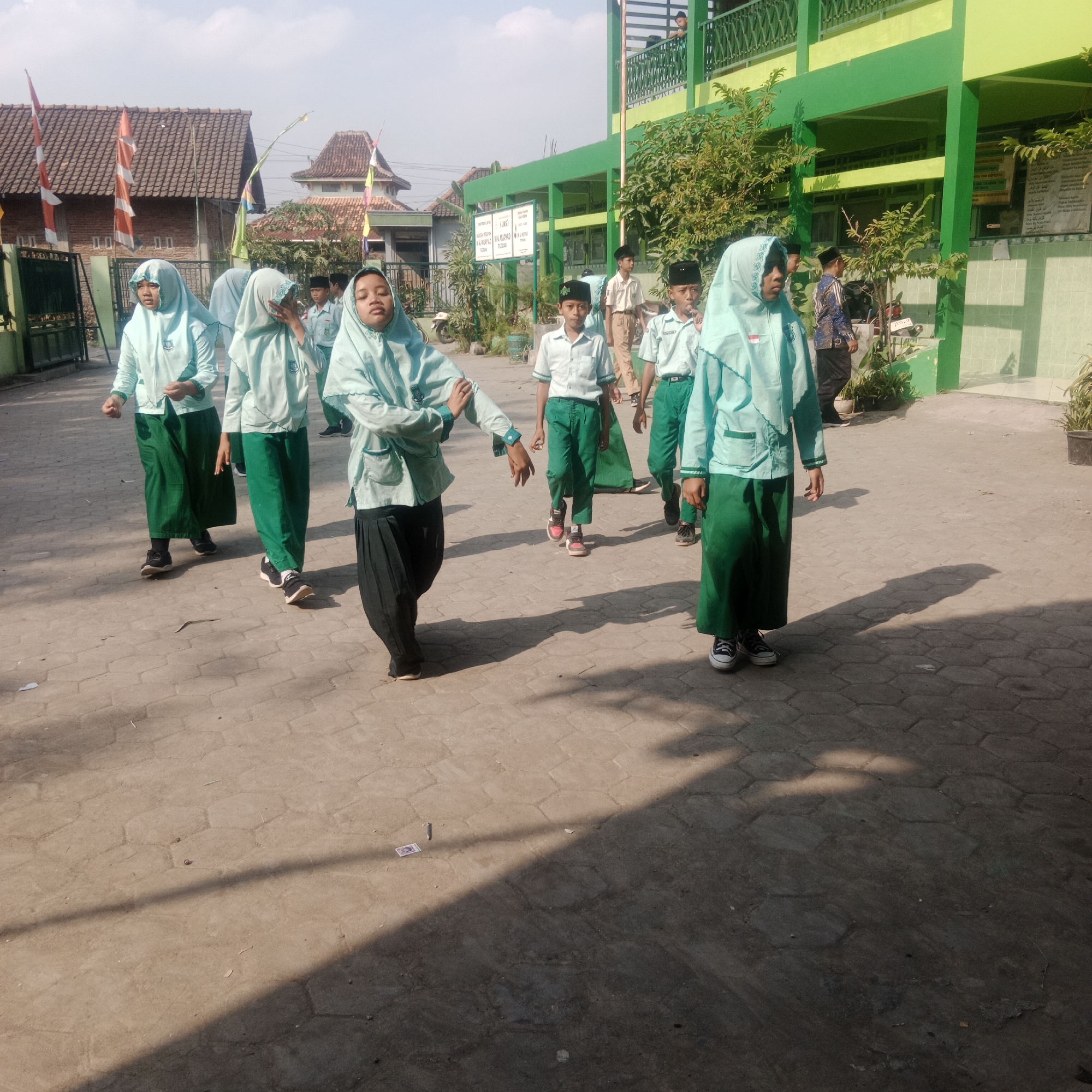 The image depicts an outdoor scene in front of a green and yellow building, likely a school, in Tegowanu Kulon, Indonesia. A group of approximately ten individuals, mostly students in light blue hijabs and green skirts or trousers, are visible walking on a paved courtyard. Some of the students are wearing white shirts and green pants. The students are arranged in a loose formation, with some walking in a line. The building behind them has multiple levels and is decorated with green trim and railings. Several trees and plants are scattered around the courtyard. The sky is clear and sunny. The ground is paved with hexagonal stone tiles. Two flags, one red and white and another white with orange and green stripes, are visible in the background. There is some debris on the ground, including what appears to be a piece of paper with an image on it.