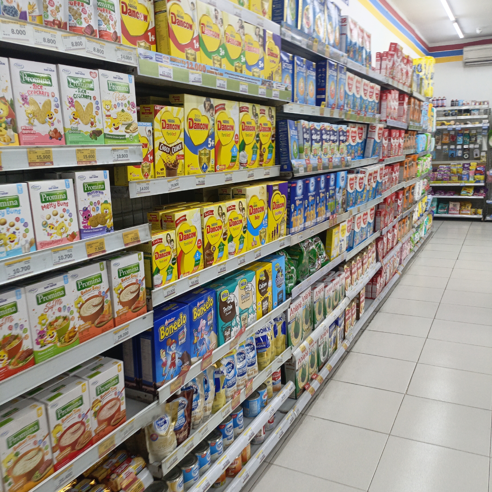 The image shows a brightly lit interior of a modern convenience store, likely a retail grocery or supermarket, with shelves stocked with various food products. The perspective is from the aisle, looking down the length of a wall of shelves. The products are predominantly packaged food items, with a focus on baby and children's food, cereals, and milk powder, displayed in colorful boxes and pouches. Prominent brands visible include "Promina" (baby food), "Nestle Dancow" (milk powder), and "Boneeto" (milk supplement). Many products are labeled with prices, such as "15.900", "10.700", "79.900", and "14.000", suggesting an Indonesian currency (Rupiah). The shelves are organized into sections, with vertical dividers. The floor is tiled with light-colored square tiles, and fluorescent lighting is visible on the ceiling, providing overall illumination. The environment is clean and organized, typical of a well-maintained retail space. There are no people visible in the frame, implying the store is either closed, or the image was taken during off-peak hours, or the people are outside the frame. The store's distinctive red, yellow, and blue striped ceiling design suggests it might be a branch of the Indomaret chain of convenience stores, a common sight in Salatiga, Indonesia.