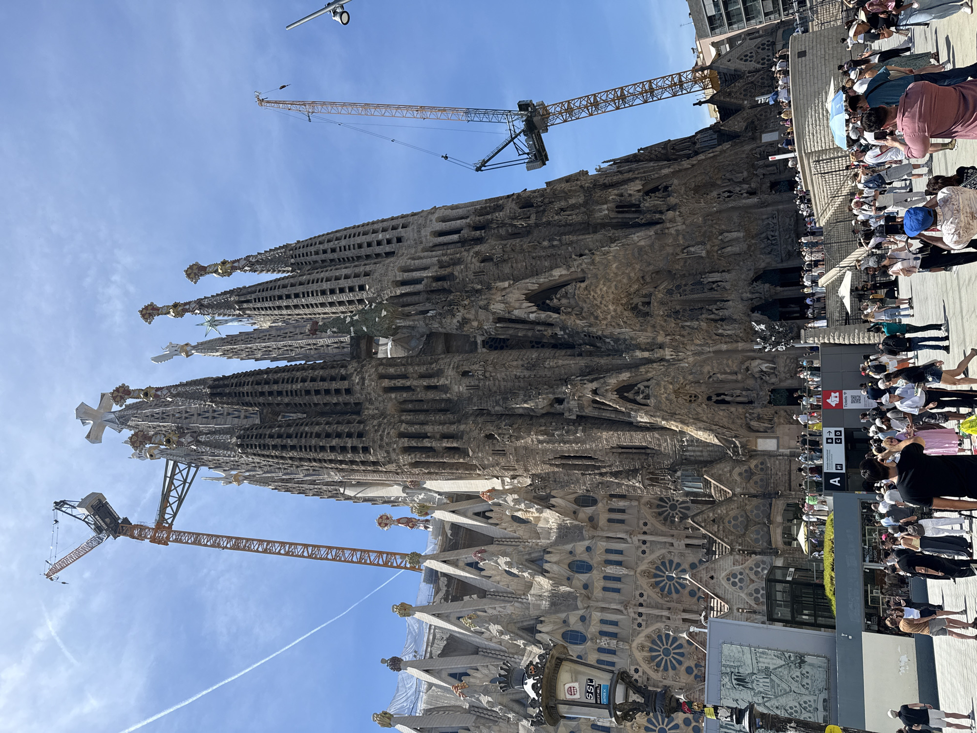 The image depicts a daytime exterior view of the Sagrada Familia basilica in Barcelona, Spain, under a clear blue sky. Two prominent construction cranes are visible, one on the left and another larger one on the right, suggesting ongoing construction or renovation work on the iconic structure. The basilica's distinctive spires, intricate facades, and detailed stonework are clearly visible.
A crowd of tourists and visitors is gathered in the foreground and around the basilica, with many taking photographs. Some individuals are standing, while others are seated. Several umbrellas are open, providing shade. A lamppost with signage is located on the left side of the image. In the background, other buildings can be seen. The overall scene captures a bustling atmosphere at a renowned landmark.