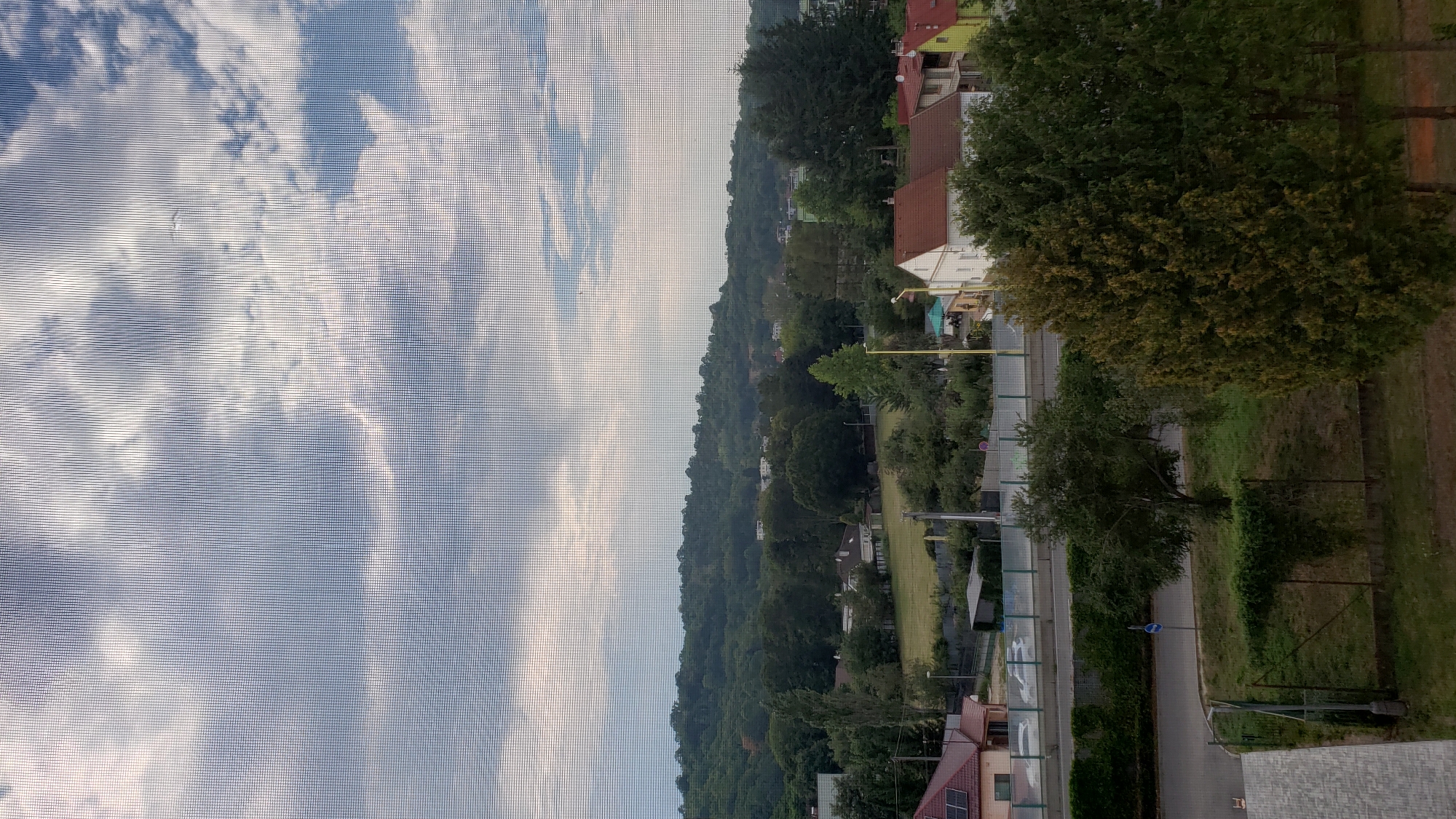 The image depicts a view from a window, looking out onto a suburban or semi-rural landscape in Prešov, Slovakia. The foreground shows a section of a road with a fence and some greenery. A small tree stands on the grass verge. Beyond the road, there are several houses with red-tiled roofs, characteristic of European architecture. Further in the background, there are more trees and a gently sloping hill covered in dense forest, suggesting the presence of a park or natural area on the outskirts of the city. The sky is partly cloudy, with a mix of blue sky and white and grey clouds. The image appears to have been taken during the daytime, likely late afternoon or early evening, given the quality of the light. There are no people or vehicles clearly visible in the immediate foreground.