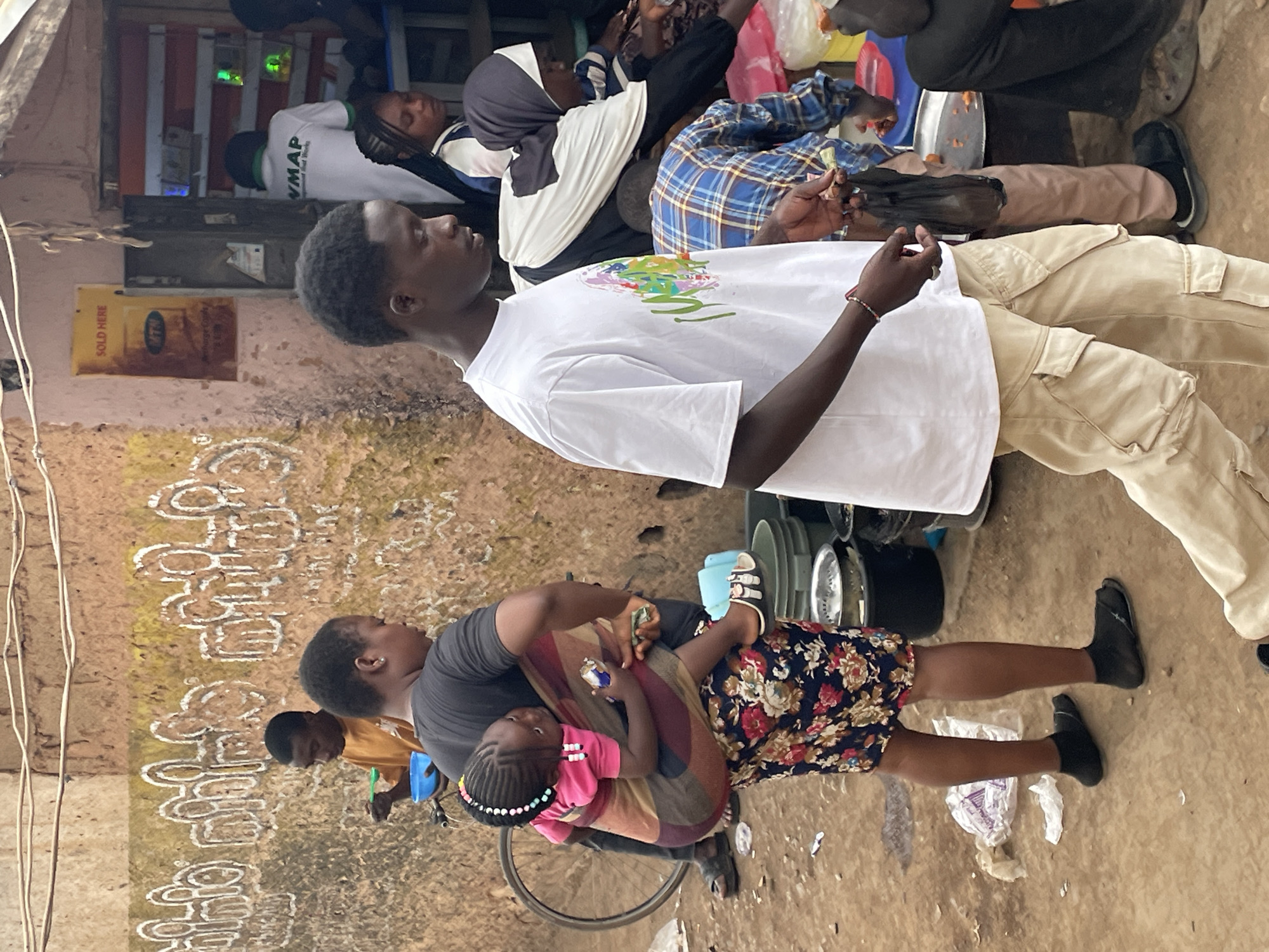 The image depicts a scene in Jos, Nigeria. A group of people are congregated outdoors. A man in a white t-shirt and khaki pants stands prominently in the foreground. Several other people are seated or lying down in the background. A woman is holding a young child, both seated on a bicycle. A wall with faded white markings is visible on the left. The ground appears to be dirt or pavement. A sign with "SOLD HERE" printed is visible in the upper left background.