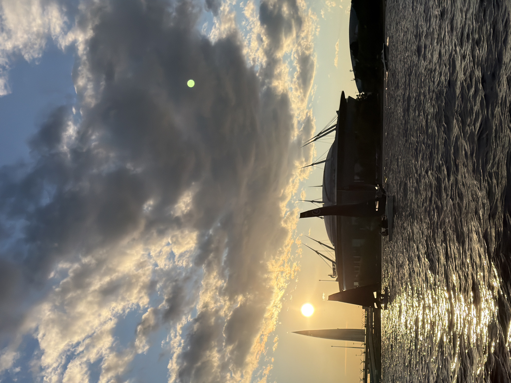 The image depicts a sunset over the Gulf of Finland in Saint Petersburg, Russia. The foreground is dominated by choppy water, reflecting the golden sunlight. Several sailboats are visible on the water, with their dark sails silhouetted against the bright sky. In the background, the distinctive architecture of Gazprom Arena, also known as the Saint Petersburg Stadium, can be seen. To the left, the Lakhta Center, a supertall skyscraper, pierces the sky. The sky itself is a dramatic mix of dark storm clouds and patches of bright, sunlit cumulus clouds, with the setting sun casting a warm glow. There is a faint, luminescent green circle in the upper right portion of the sky, possibly a lens flare or artifact. The overall scene captures a moment of dynamic weather and iconic urban landmarks.