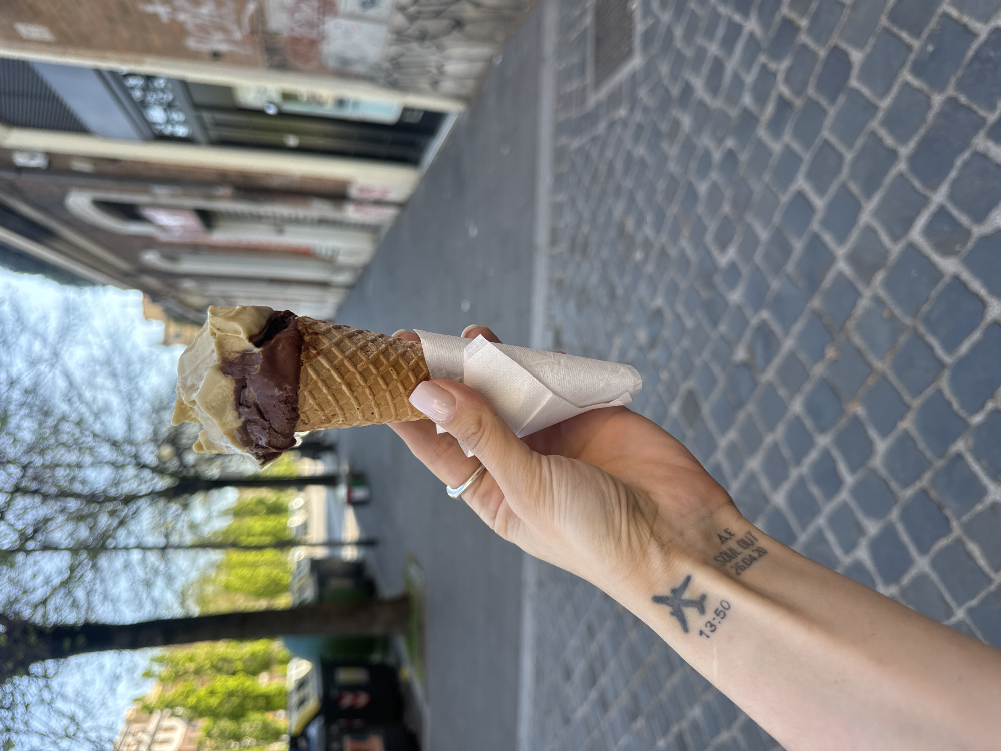 A woman's hand holds an ice cream cone in front of a cobblestone street and building. The ice cream cone is wrapped with a white napkin and contains two flavors of ice cream, light-colored and dark-colored. The woman's fair-skinned hand is adorned with a silver ring and a tattoo depicting a plane, time, and a phrase "At Soul Out 26.04.20". The background shows a street in Rome, Italy, with buildings and trees on either side.