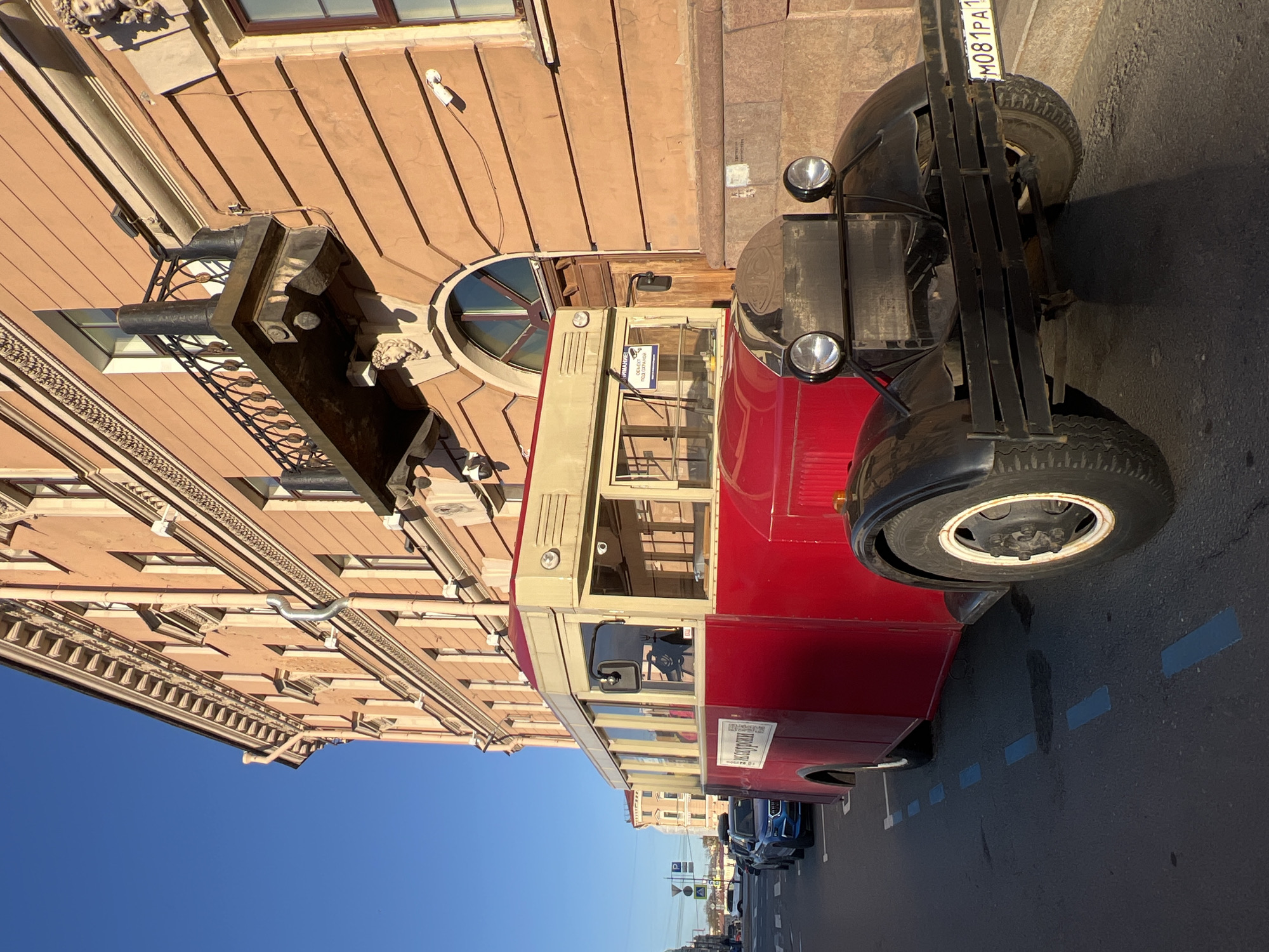 A red, vintage passenger bus is parked on a city street next to a tan-colored building. The bus has black fenders and a black front grille. A Russian license plate is visible on the front of the vehicle. The building is ornate, with architectural details such as a balcony and decorative moldings. The sky is a clear blue. Based on the architecture and the presence of Russian text, the location is likely in Saint Petersburg, Russia.