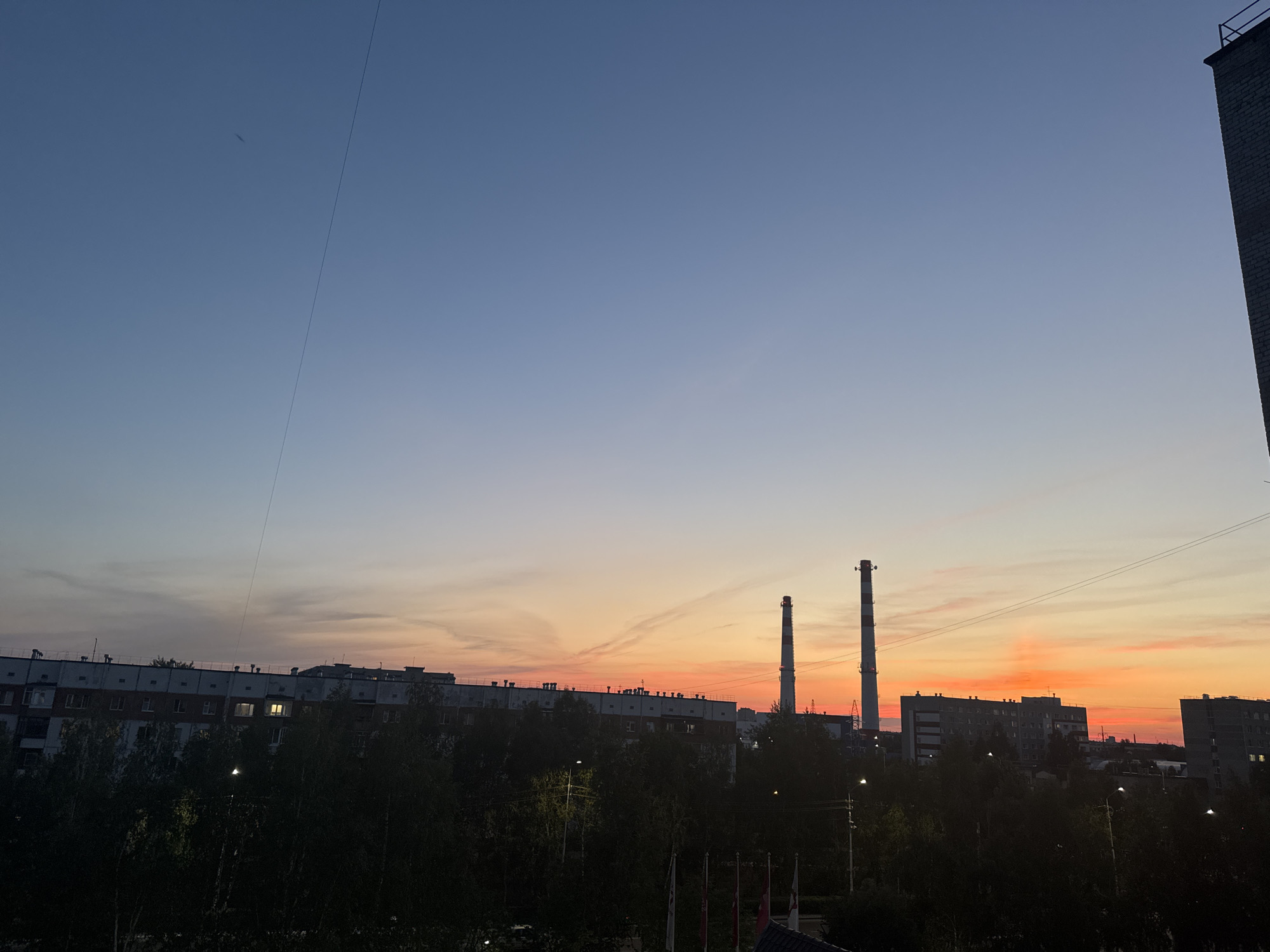 The image depicts a sunset scene in Surgut, Russia. The sky displays a gradient of colors, transitioning from a deep blue at the top to vibrant oranges and pinks near the horizon. Silhouetted against the colorful sky are industrial chimneys, part of a power plant, and the outlines of apartment buildings and trees. The overall impression is of an urban industrial landscape at dusk.