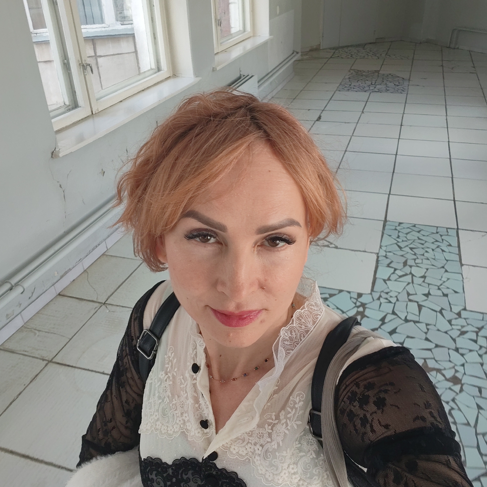 A woman with short, light reddish-brown hair is taking a selfie in what appears to be a public building in the city of Chaikovsky, Russia. She has fair skin, dark eyebrows, and eyelashes, and is wearing lipstick. The woman is wearing a white blouse with black lace sleeves and a black lace trim along the chest. She also has a necklace on. A black backpack with leather straps is visible on her shoulders. The background is a hallway with white-tiled walls and windows on the left side of the frame. There are old radiators below the windows. The floor has a pattern of white tiles interspersed with sections of blue mosaic tiles. The hallway appears somewhat worn, with cracks visible in the walls and floor.