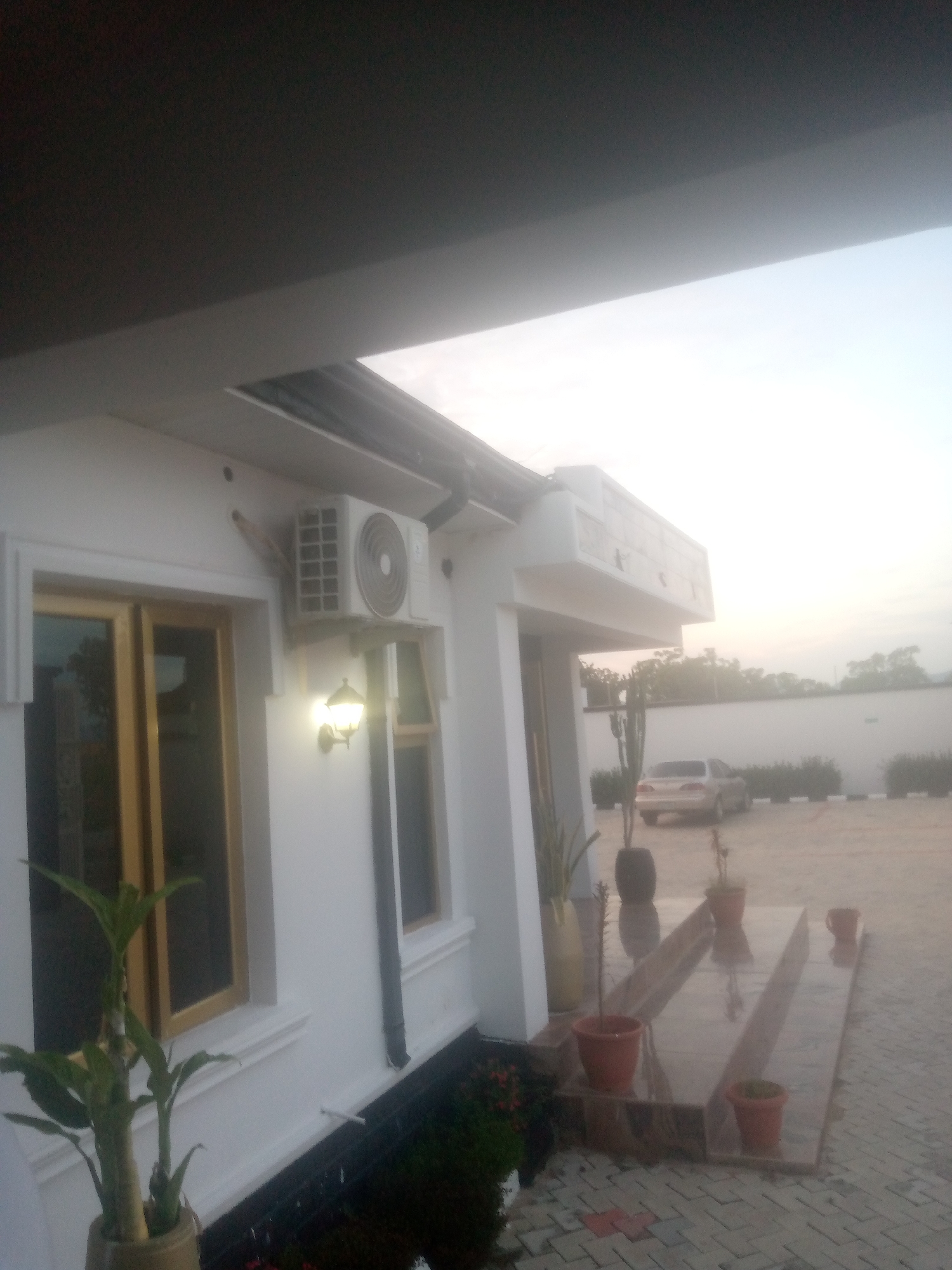 In Jauro Yinu, Nigeria, a white building with wooden-framed windows is visible. An air conditioning unit is mounted on the exterior wall, positioned above an illuminated wall-mounted lamp. A paved walkway leads to a set of steps, bordered by potted plants. A light-colored car is parked in the background, with a white wall and a line of green bushes behind it. The sky is overcast, suggesting either dawn or dusk. The image appears to be taken from a low angle, looking up towards the building's eaves.