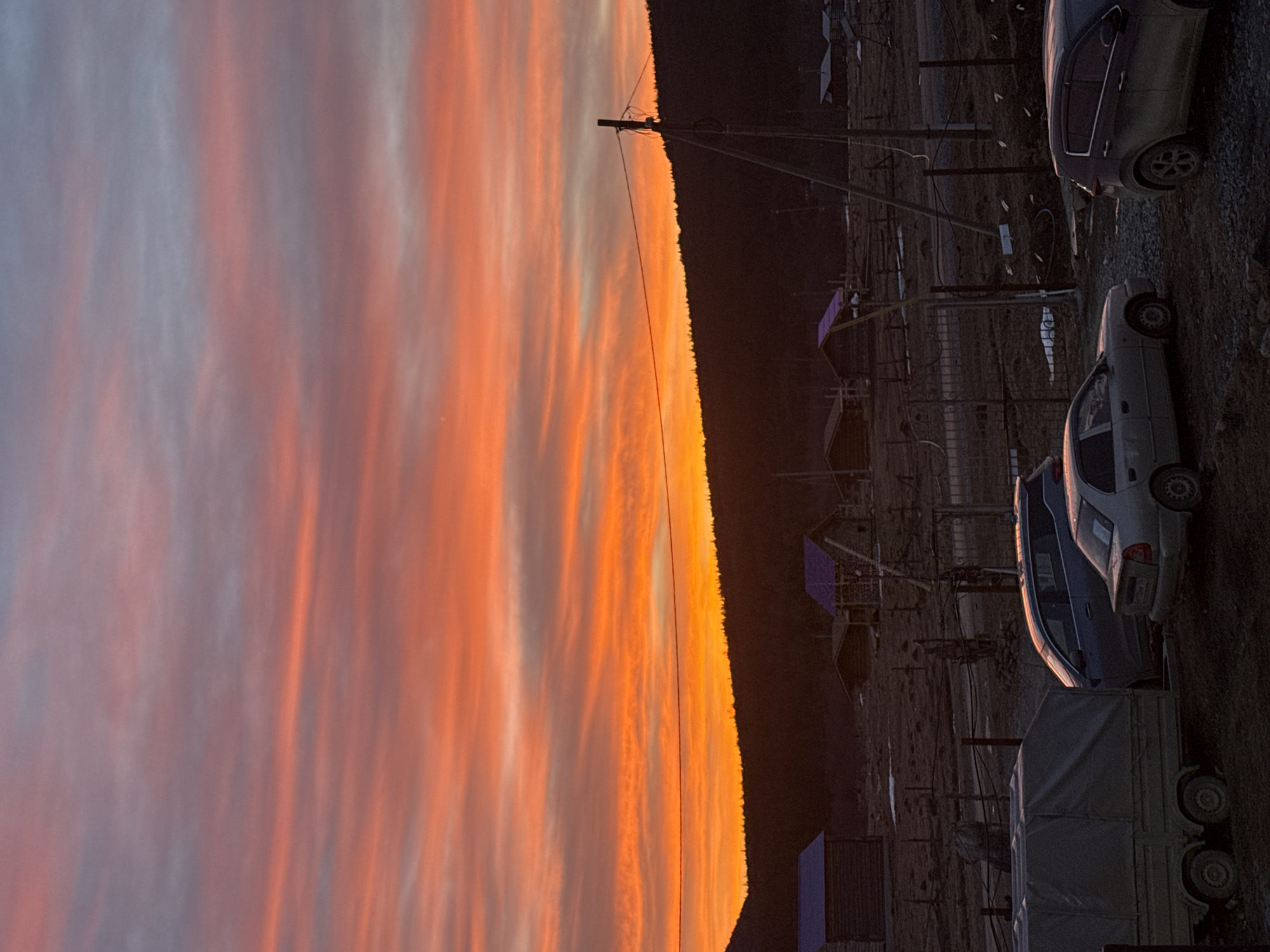 The scene depicts a rural area in Tyulyuk, Russia, at either sunrise or sunset. The upper portion of the frame is dominated by a sky displaying horizontal bands of vibrant orange, red, and pink clouds, transitioning to lighter pink and grey-blue at the zenith. The lower portion of the sky is obscured by a dark, silhouetted forested hillside or mountain range, forming the horizon.
In the midground, several rustic, dark-stained wooden cabins or houses are visible, some featuring brightly colored roofs (blue and purple). These structures are sparsely distributed across an uneven, barren terrain with visible patches of snow or ice. Fences constructed from wire and wooden posts delineate portions of the landscape. A tall wooden utility pole with attached power lines stands prominently on the right side of the midground.
The foreground consists of a dirt and gravel area serving as a parking surface. Parked vehicles include, from left to right: a light-colored two-axle utility trailer with a grey canvas cover; a blue-grey sedan; a light silver sedan, possibly a Hyundai Accent, with a visible Russian license plate "K 550 KK 96" on its rear; and a dark-colored SUV or hatchback, possibly a Kia Sportage, exhibiting accumulated dirt on its lower body panels and wheels. No individuals are visible within the frame, and no discernible actions are in progress.
