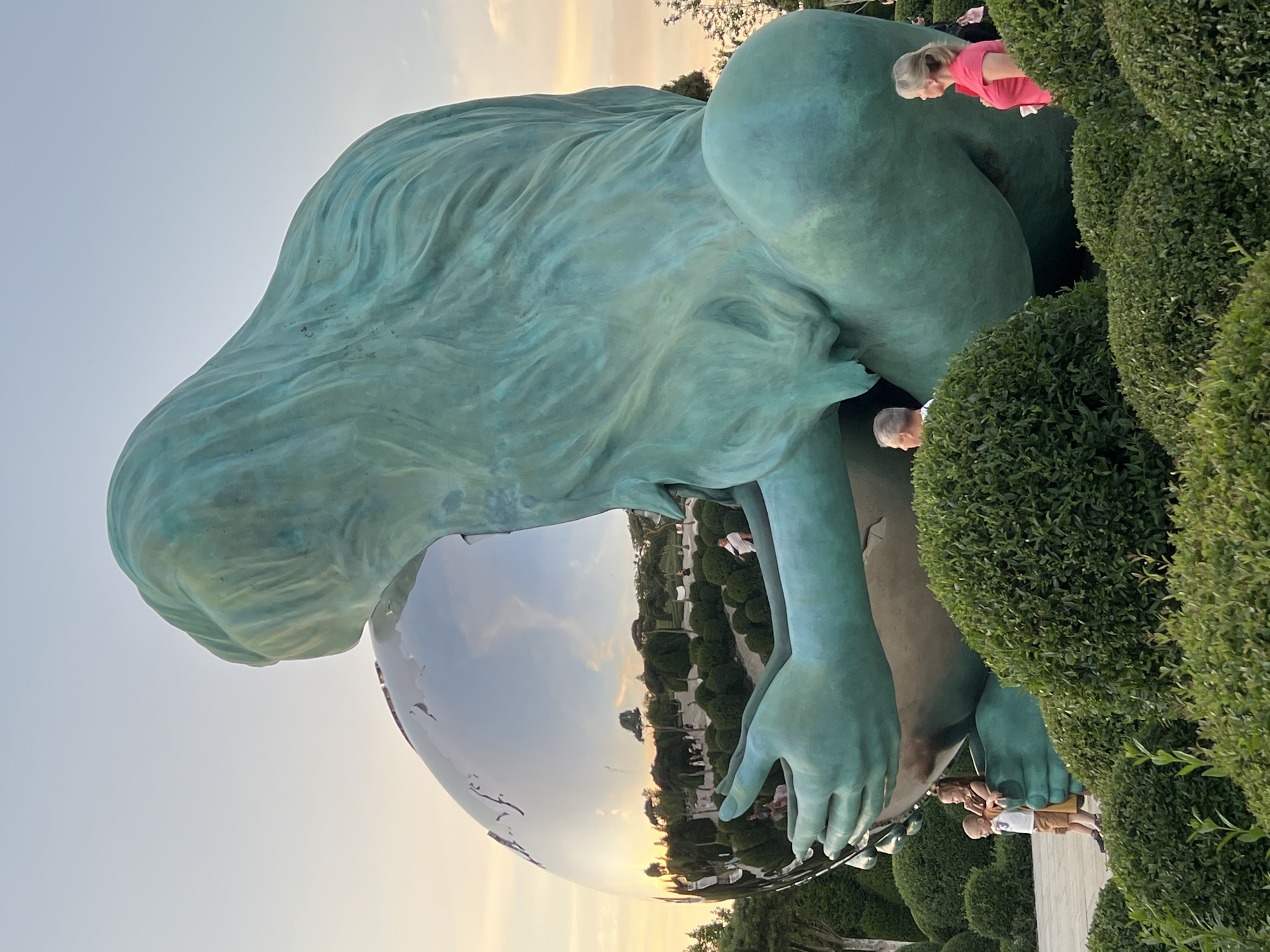 In the city of Krasnodar, Russia, a large, teal-colored statue of a mythical female figure cradles a giant, reflective sphere. The statue's long, flowing hair cascades down its back, and its hands gently cup the sphere. The sphere reflects a park-like environment with manicured bushes, trees, and a sunset sky with scattered clouds. Several people are visible in the background, observing the sculpture and the surroundings. Among them, two children stand near the base of the statue, while a man and a woman are seen further back, looking towards the sculpture. The scene is bathed in the warm light of the setting sun.