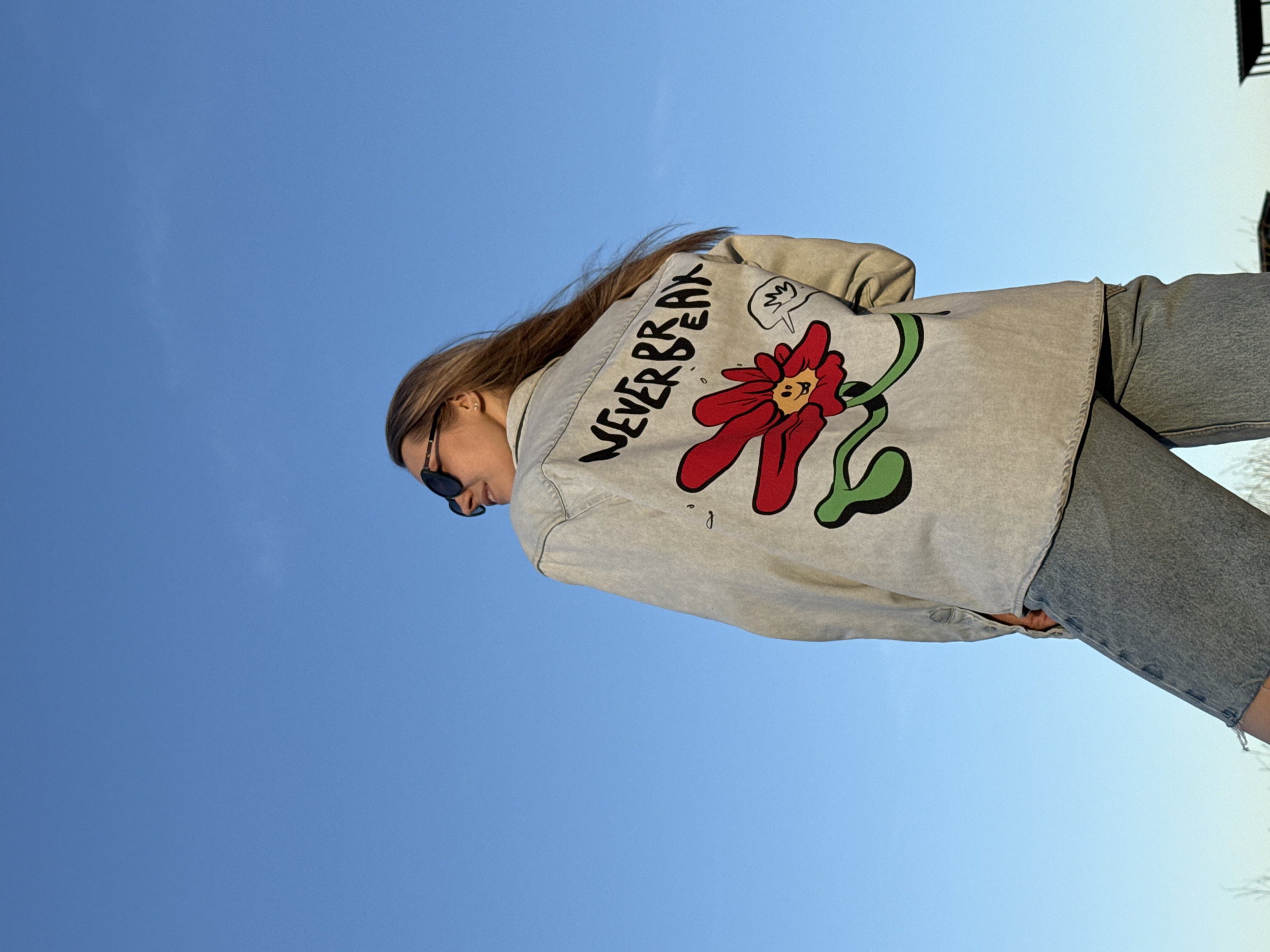 A young woman stands outdoors under a clear blue sky, facing away from the camera. She is wearing sunglasses, light grey jeans, and a grey denim jacket with a large stylized floral design on the back. The floral design features a red flower with a cartoonish face and green stems. The words "NEVER BREAK" are written in black above the flower. The location is Chaikovsky, Russia.
