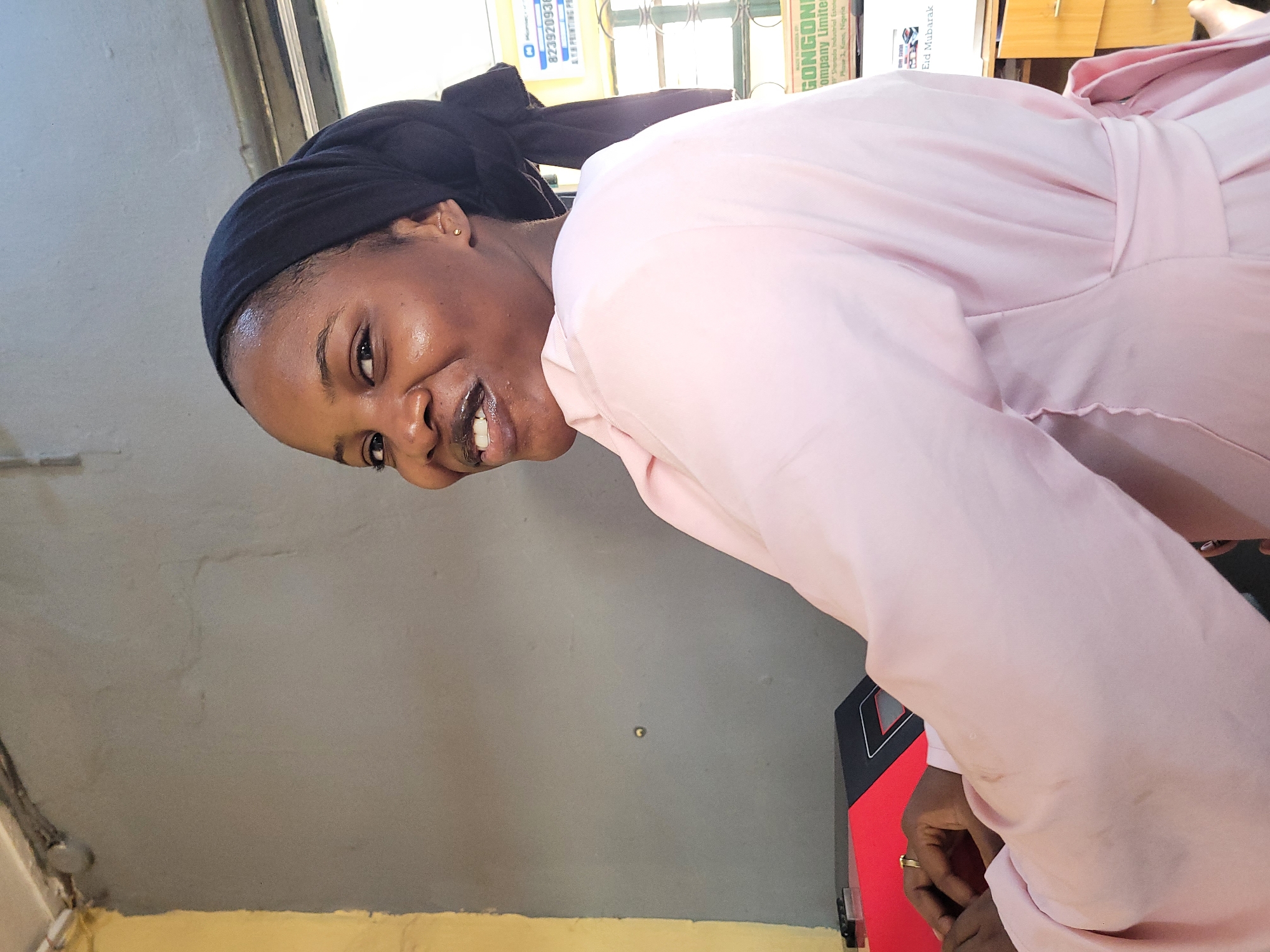 A woman with dark skin and wearing a black head covering smiles at the camera while wearing a pink robe-like garment. The background is a mix of a gray wall and what appears to be an office setting. On a shelf or desk are boxes with labels that include "GONGON Company Limited," "Eid Mubarak," and a partial phone number "823920938." The location information from the label suggests the image was taken in or near Kano, Nigeria, not Jos.