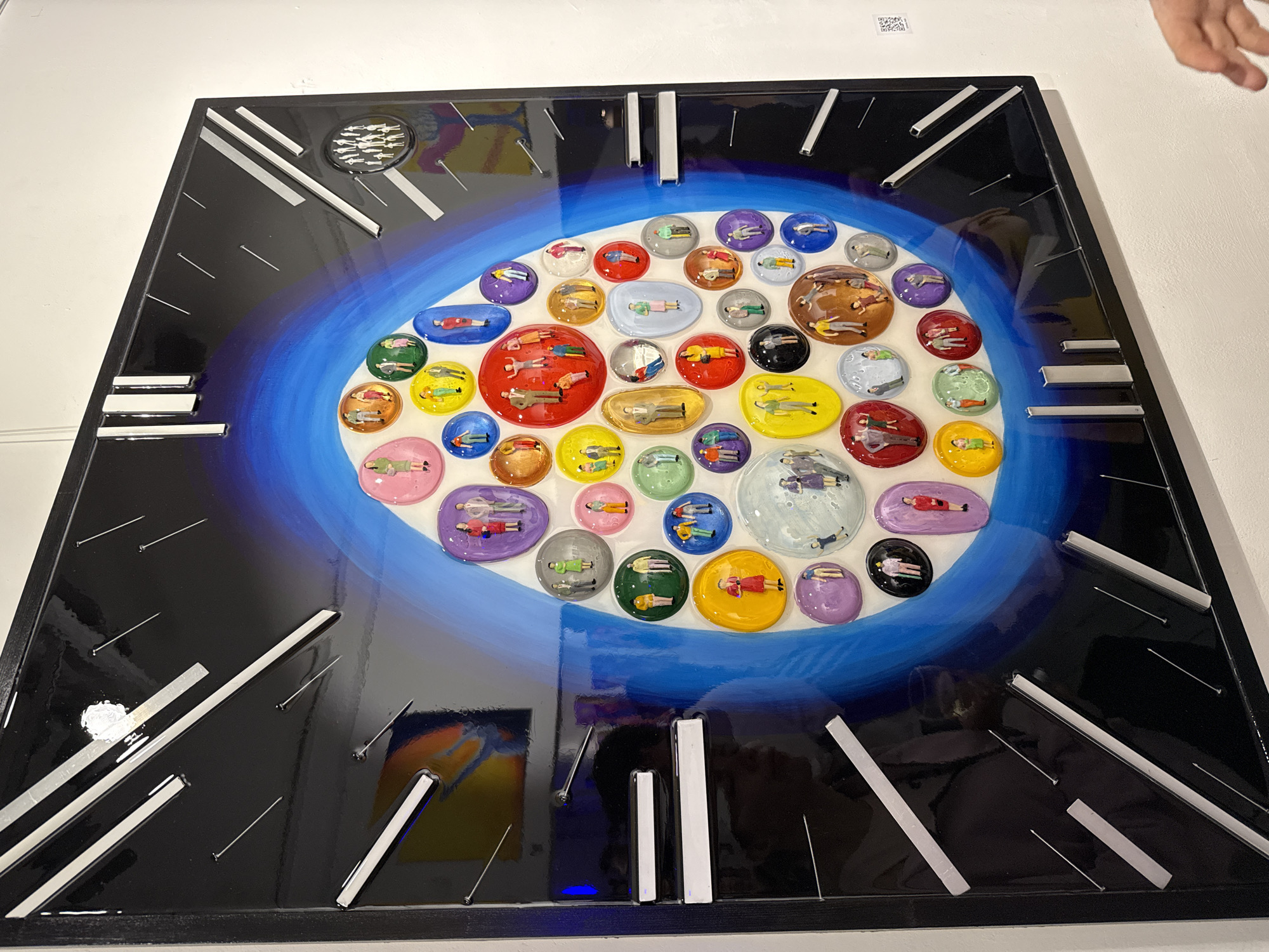 The image shows a square artwork in a black frame, likely hanging in a gallery or exhibition space. The artwork features a glossy black background with twelve silver geometric markers arranged around the perimeter, reminiscent of clock face numerals. A gradient of blue paint creates an oval shape at the center. Within the oval, numerous small, colorful, glossy convex circles or ovals are clustered together. Each circle contains a tiny figure of a person, depicted lying down. The overall impression is of a clock or symbolic representation of time with individuals contained within moments or periods. The location in Moscow, Russia is not determinable.