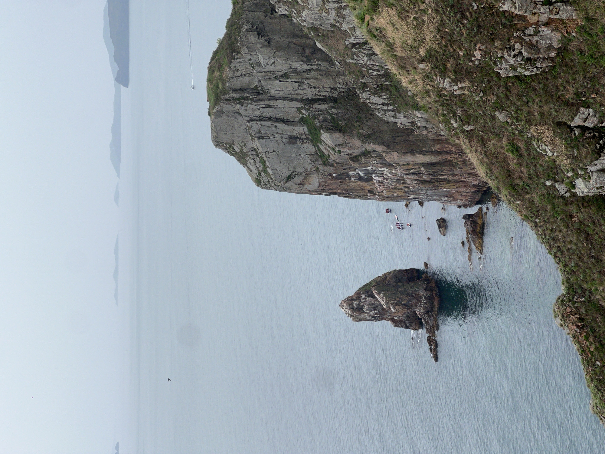 The image shows a high-angle view of a rocky coastline in Russkiy, Russia. A large, steep cliff face dominates the right side of the frame, covered in vegetation. To the left of the cliff, a smaller rocky outcrop rises from the water. Several boats are visible near the base of the cliff, and other landmasses can be seen in the distance through the overcast sky. The sea is calm and stretches out to the horizon.
