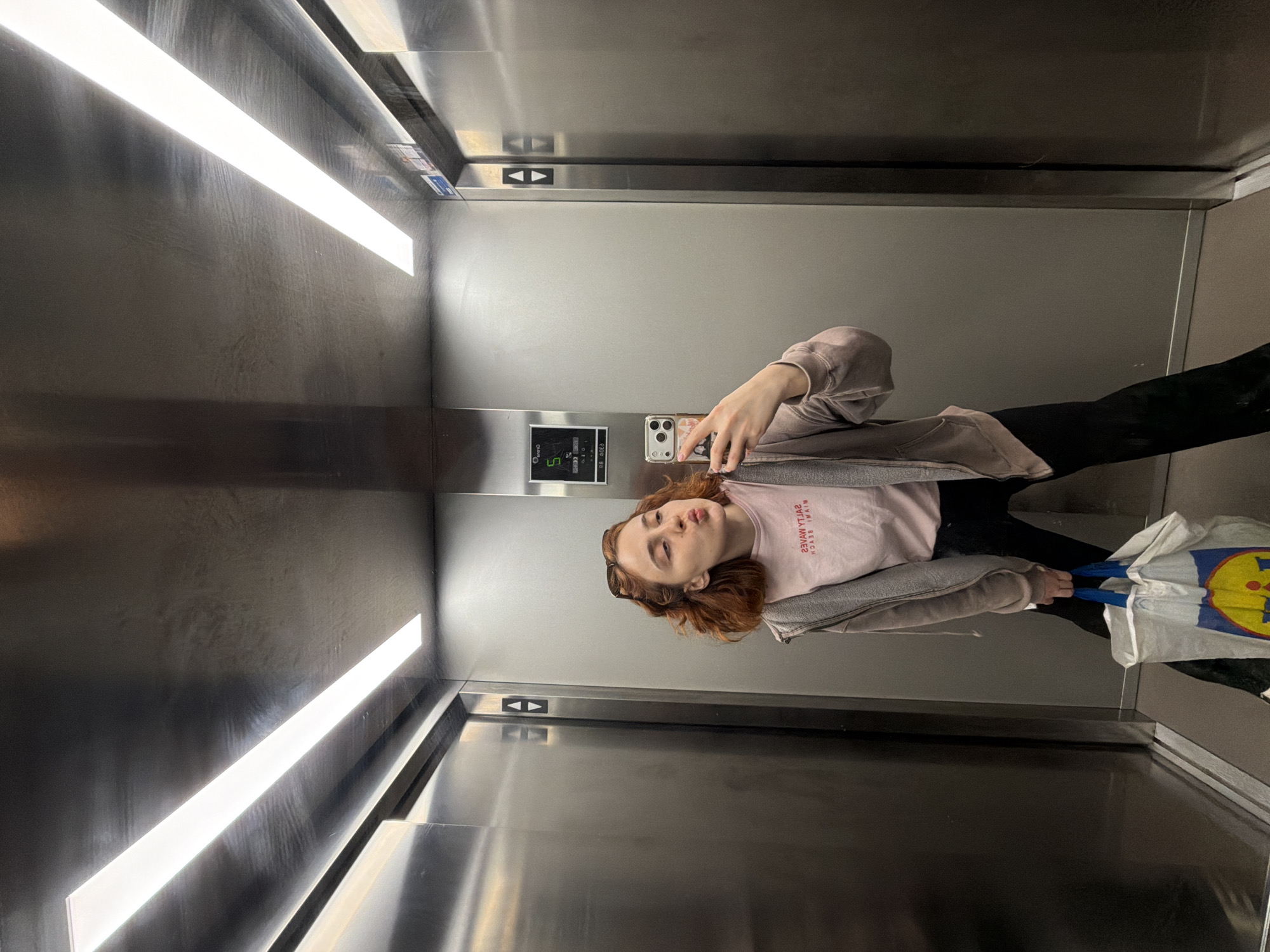 A young woman with reddish-brown hair is taking a selfie in an elevator. She is wearing a light pink t-shirt with red text that reads "SALTY WAVES MIAMI BEACH" and a light gray zip-up hoodie. She has black pants on and is holding a reusable shopping bag with the Lidl logo. Her mouth is puckered as if she is about to kiss. The elevator has stainless steel walls and two long fluorescent lights on the ceiling. There are elevator buttons visible on the wall behind her, and the elevator car number "5" is also displayed. The scene is not identifiable as being in the city of Strasbourg, France.
