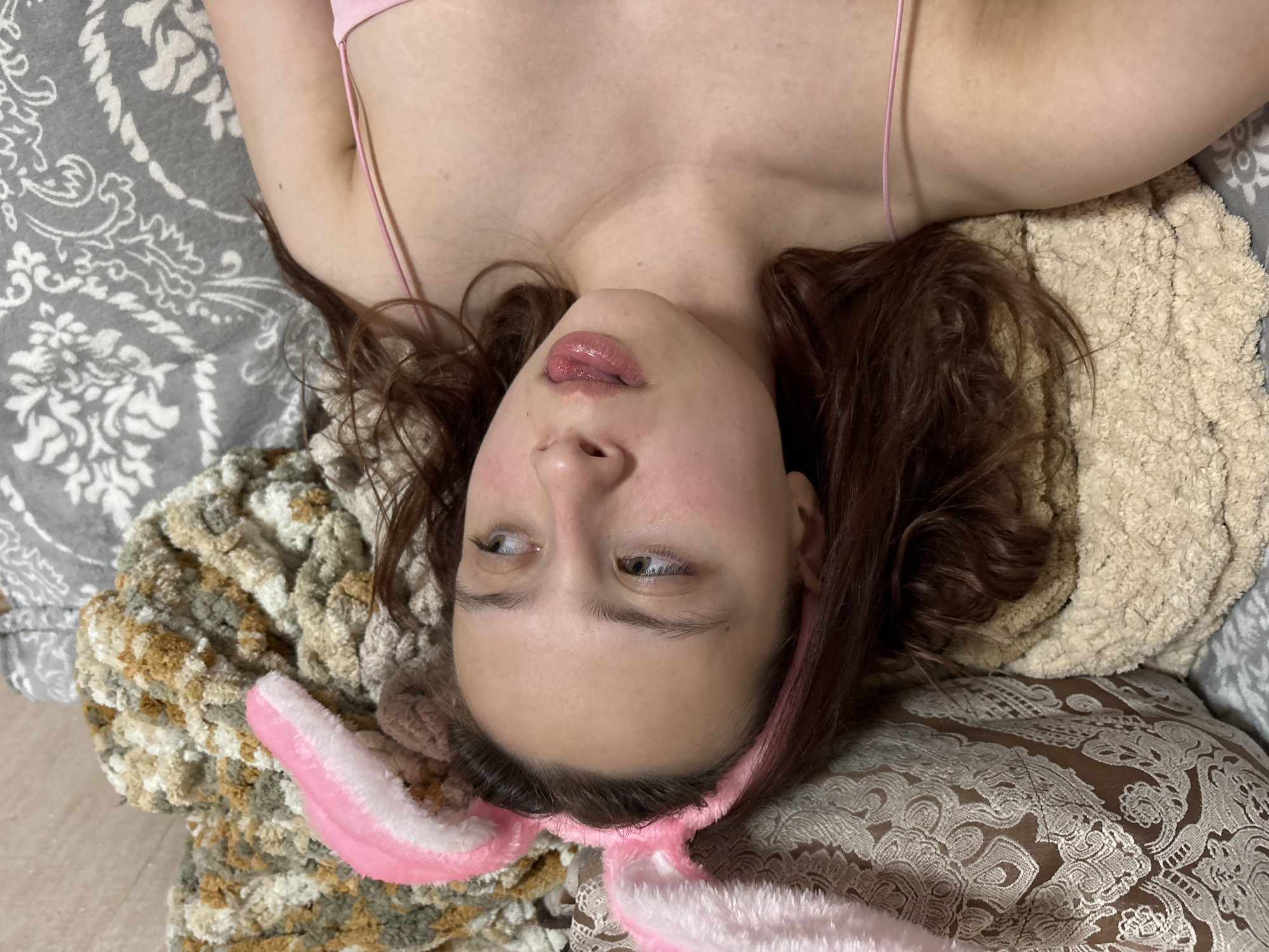 An overhead, close-up shot captures a person, likely female, lying on their back in a relaxed indoor setting. The person's head is positioned towards the bottom of the frame, adorned with a fluffy pink and white bunny ear headband. Their long, wavy reddish-brown hair is spread around their head, resting on various soft textures.
The person has fair skin, and their eyes are cast slightly to the side, conveying a contemplative or perhaps playful expression. Their lips are slightly parted and appear to have a glossy finish. They are wearing a simple pink top, visible as a thin strap over the shoulder and across the upper chest.
The person is surrounded by an assortment of soft furnishings. To the left and upper side of the frame, there is a gray and white blanket or pillow featuring an intricate damask-like pattern. Below the person's head, a chunky, textured knitted or crocheted blanket in shades of beige, white, and brown is visible. To the right, another fluffy, light beige or tan blanket provides cushioning.
The scene suggests a relaxed atmosphere, likely in a bedroom or living area. No specific activity beyond lying down is clearly depicted, though the pose and bunny ears hint at a playful or leisure moment. There is no visible text in the image. The time of day and external weather are not ascertainable from the indoor setting, and no visual cues within the image confirm the provided location context of Прикамский, Russia.