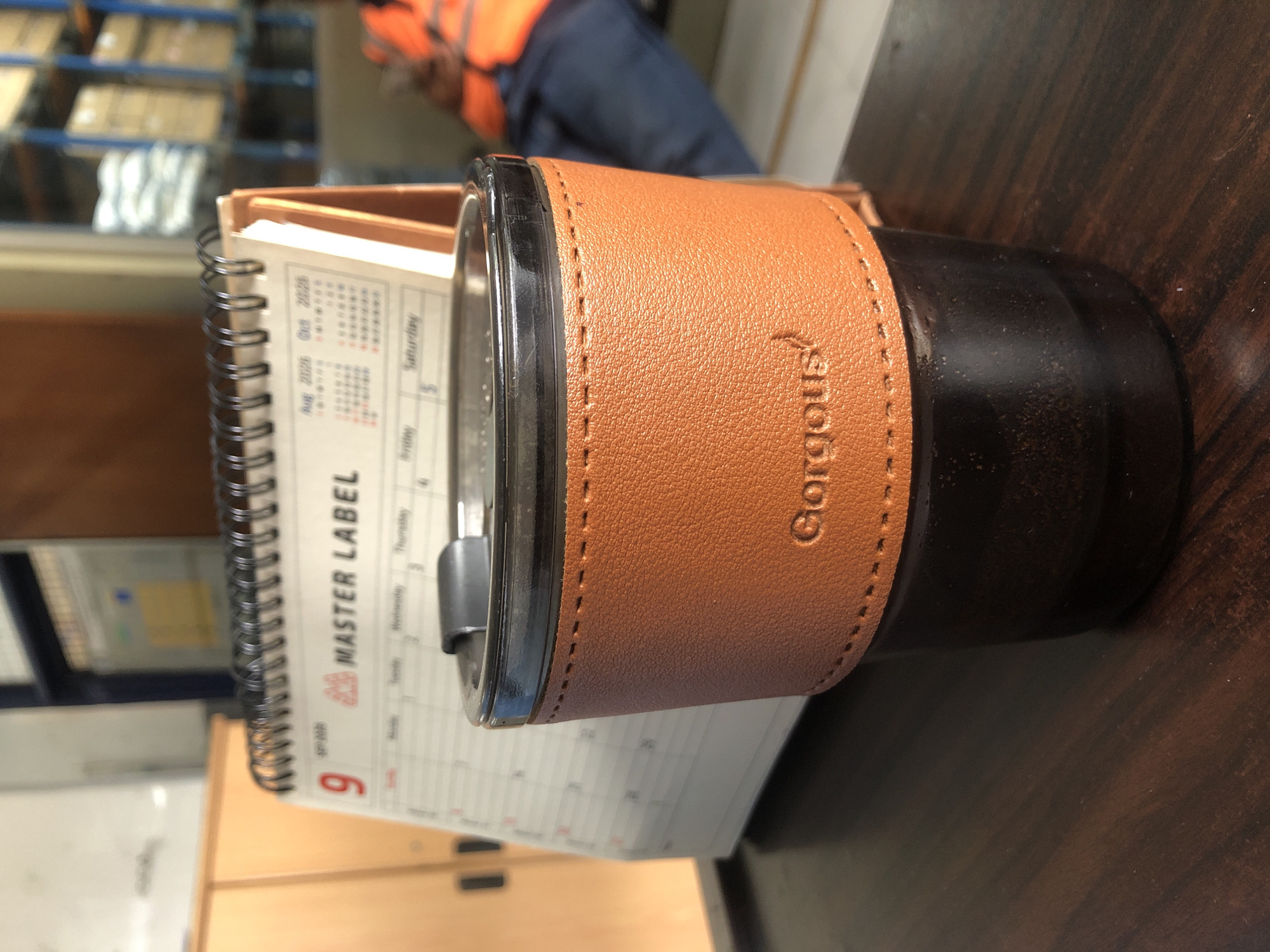 A brown, leather-sleeved tumbler sits on a dark wooden desk in front of a desk calendar displaying "MASTER LABEL" and the month of September. The tumbler has a black lid and a stitched leather sleeve embossed with the word "Gorgous." In the background, shelves with boxes are visible, and a person wearing an orange vest is partially visible. The scene appears to be in a warehouse or office environment.