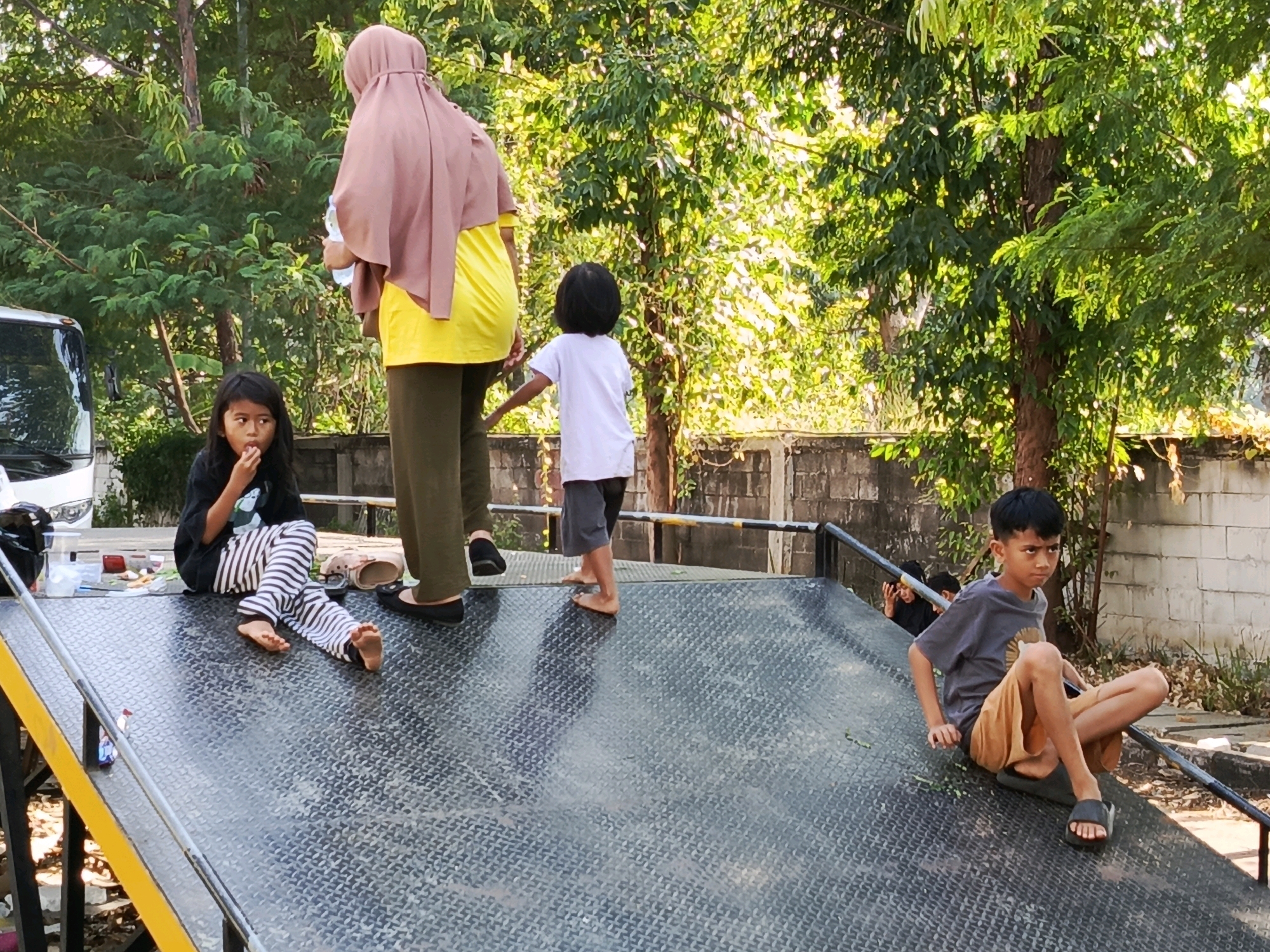 A group of people, including two young girls, a young boy, and two adults, are gathered on a metal ramp outdoors. The girl on the left is sitting down, wearing a black t-shirt with a graphic and striped pants, and appears to be eating. The boy on the right is sitting on the ramp, wearing a gray t-shirt and brown shorts, and is looking towards the camera. The adult on the left is wearing a pink headscarf and a yellow shirt. The ramp is textured with a diamond plate pattern and has yellow and black railings. In the background, trees and a wall are visible. The scene appears to be located in a suburban or residential area in Tangerang, Indonesia, possibly a park or a community space.
