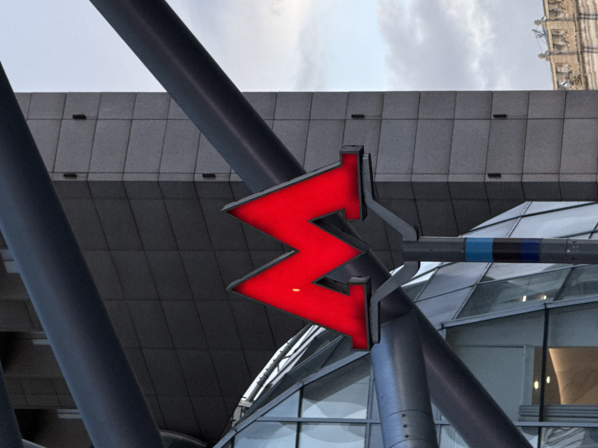 This is a medium shot of the illuminated red "M" symbol of the Moscow Metro. The symbol is mounted on a grey pole with blue and black stripes. The background consists of a modern building with a grey tiled facade and large glass windows, as well as a cloudy sky. The composition is framed by thick, dark grey pipes that intersect the image. The lighting suggests it is either dusk or an overcast day. The photograph appears to have been taken in Moscow, Russia, given the iconic metro symbol. There are no people visible in the image.