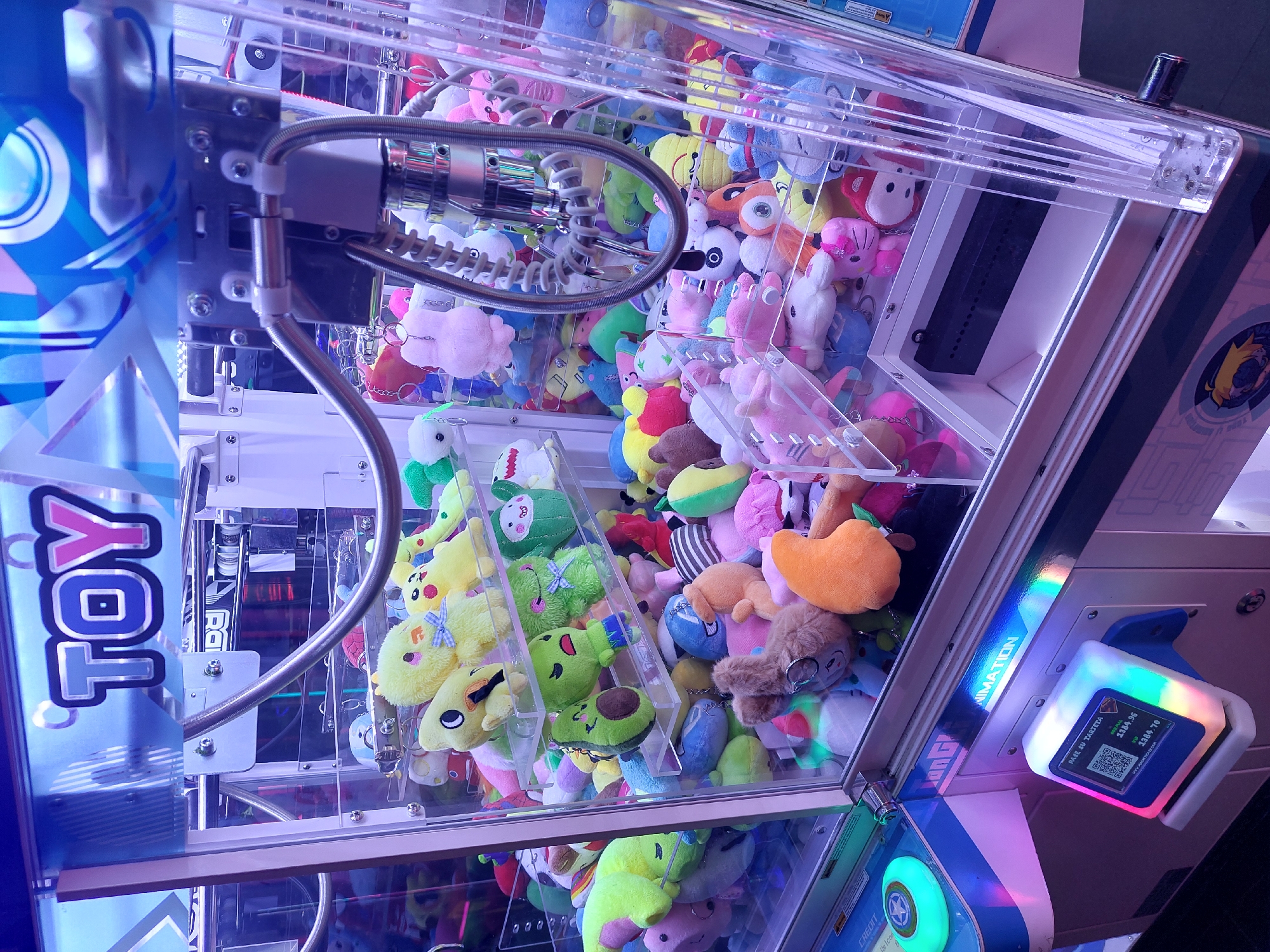 A brightly lit claw machine, filled with numerous plush toys, is captured from a slightly elevated perspective. The transparent acrylic casing allows a clear view of the diverse collection of stuffed animals packed tightly within. Among the plushies are various cartoon characters and animal shapes, including yellow figures resembling Pikachu, green avocado-shaped toys, pink bunnies, and other colorful creatures. The metallic claw mechanism, with its coiled cable, is visible at the top of the machine, positioned above the pile of toys.
On the left side of the machine, a blue panel features the word "TOY" written vertically in white with a pink outline. The machine's internal mechanics and metal framework are discernible through the transparent panels. On the right side, a payment terminal or card reader glows with vibrant rainbow colors. Text on this terminal includes "PASE SU TARJETA" (Pass your card), a QR code, and monetary values such as "$249.90" and "$384.70," along with a partially visible website "WW.PLAYTOTEM.COM". The word "ANIMATION" is also visible horizontally on the side panel of the machine. Below the card reader, a circular, glowing blue and green button or control is present. The machine appears to be in an arcade or entertainment venue, ready for a player. The visible Spanish text on the payment terminal aligns with the provided context of Buenos Aires, Argentina.