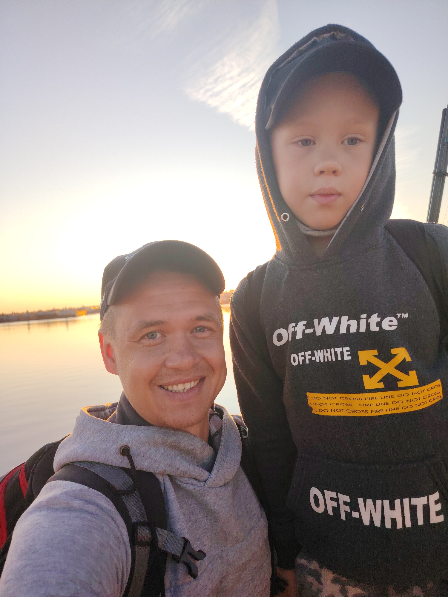 The image shows two people, an adult male and a young boy, standing in front of a body of water at sunset. The adult male, on the left, is wearing a grey hoodie and a black baseball cap. He is smiling and looking at the camera. The young boy, on the right, is wearing a black hooded sweatshirt with "Off-White" branding and a black baseball cap underneath the hood. He is looking directly at the camera. The background shows a body of water with a hazy sunset casting a warm glow. In the distance, there appears to be a cityscape or industrial area along the shoreline. This scene is consistent with the landscape of Chaikovsky, Russia, which is located on the Kama River and known for its industrial presence.