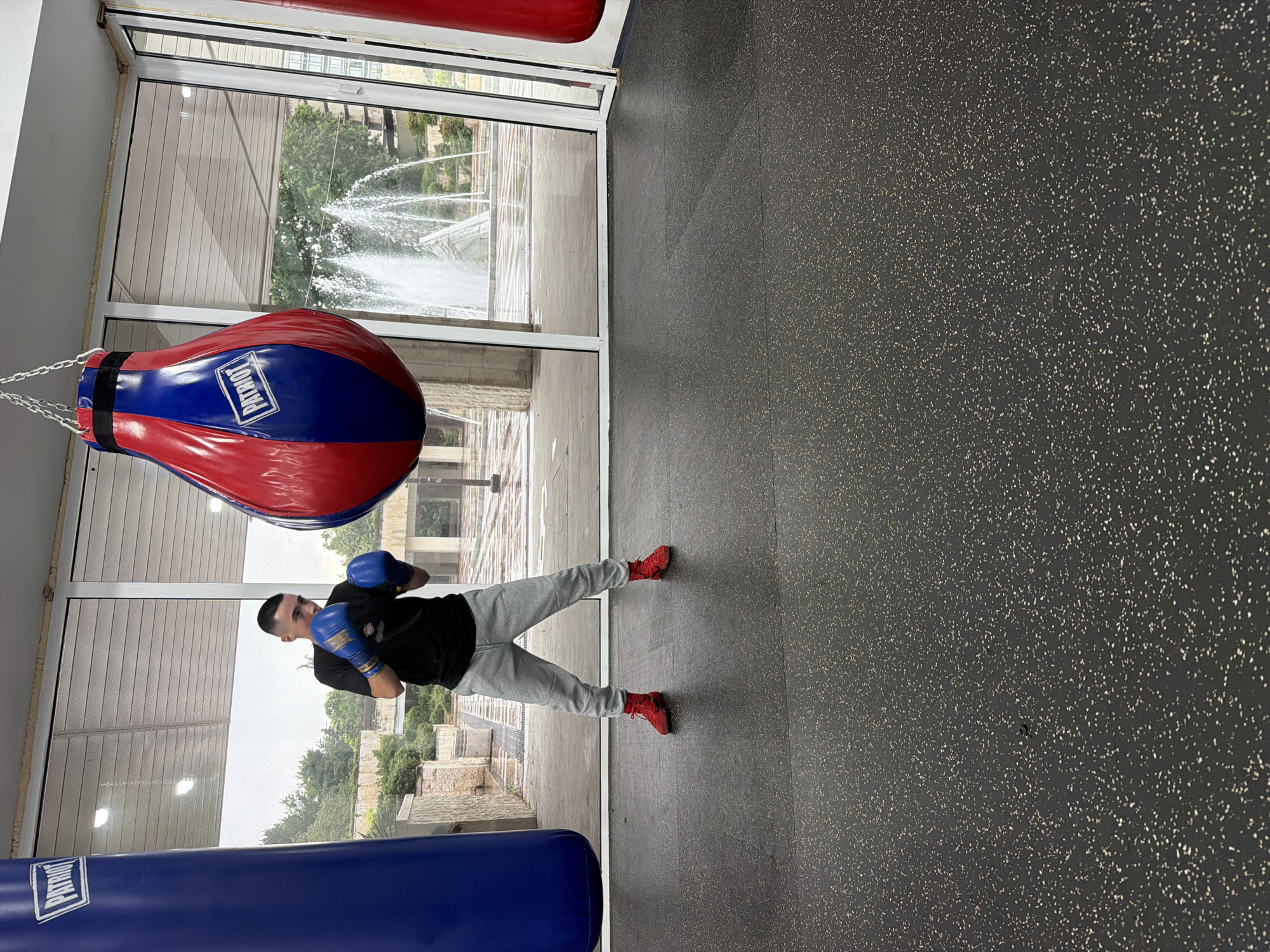 A young man is depicted in a boxing gym, positioned in a focused stance while wearing blue boxing gloves, a black t-shirt, grey sweatpants, and red athletic shoes. He is situated on a dark grey, speckled rubber gym floor. To his left, a red and blue tear-drop shaped speed bag hangs from the ceiling by chains, displaying the word "PATRIOT" in white lettering on its blue section. A larger, blue cylindrical punching bag, also bearing the "PATRIOT" logo, is partially visible in the foreground.
Large floor-to-ceiling windows behind the man offer a view of an outdoor urban environment. Outside, a prominent fountain with multiple water jets sprays upwards, surrounded by trees and architectural structures. The bright lighting suggests it is daytime with clear weather. The overall scene captures an athlete training indoors with a glimpse of the city of Blagoevgrad, Bulgaria, outside. The man's expression is one of concentration.