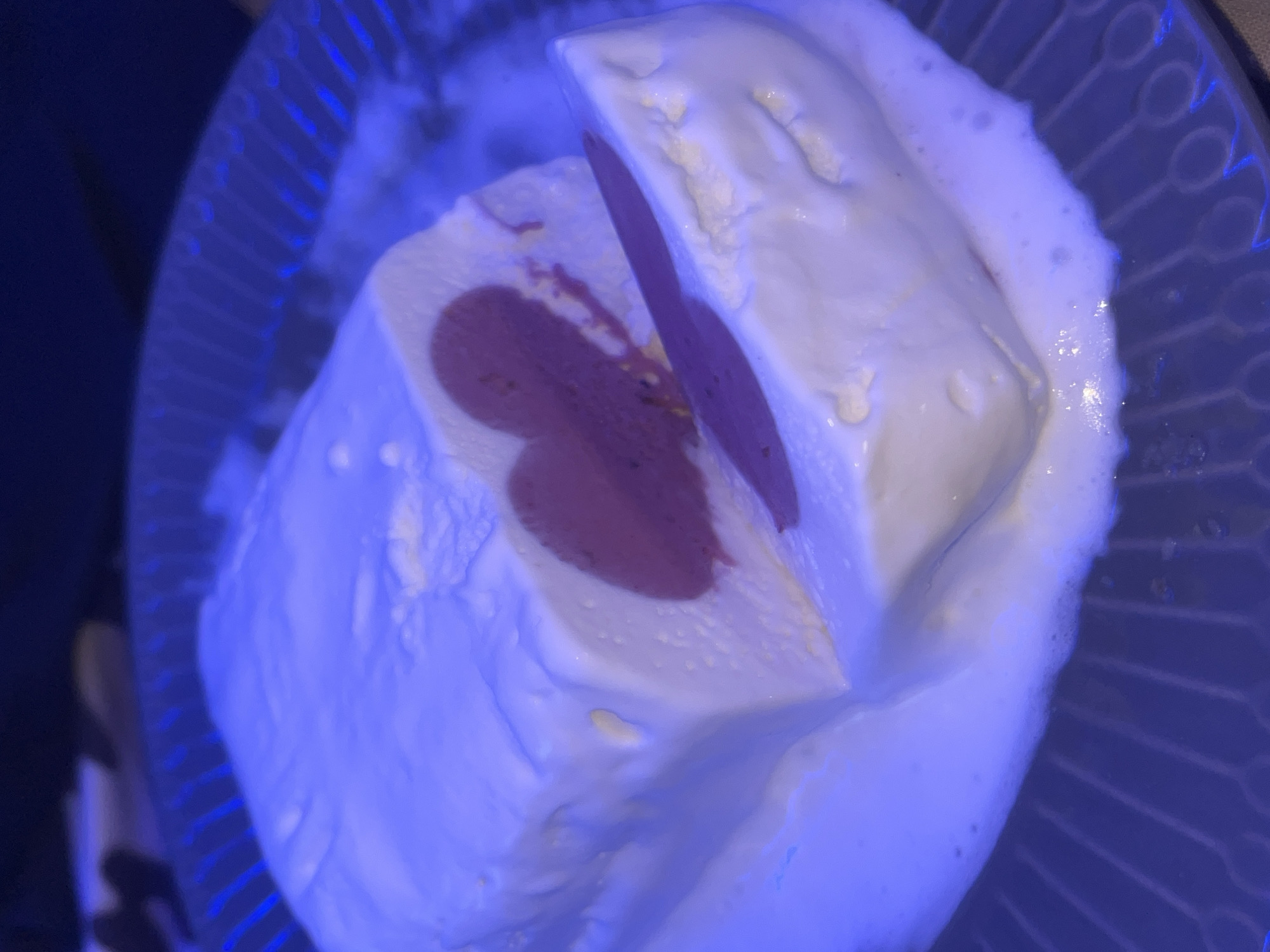 The image displays a close-up of a dessert, likely an ice cream cake or frozen mousse, served on a clear, patterned plate. The dessert features a white outer layer, possibly vanilla or whipped cream, encasing a distinct, heart-shaped core of a deep reddish-purple or maroon color, suggestive of a fruit-based component like sorbet or jam. A slice has been removed from the dessert, revealing its internal structure.
Around the base of the dessert, there are indications of slight melting, with some foamy liquid visible on the plate. The transparent plate has a decorative radial or sunburst pattern. The entire scene is bathed in a prominent blue light, which casts a cool, artificial hue over all elements and highlights the plate's design. The background is dark and out of focus. No people or text are visible in the image. The strong blue lighting suggests an indoor setting, possibly during the evening or under specific mood lighting.