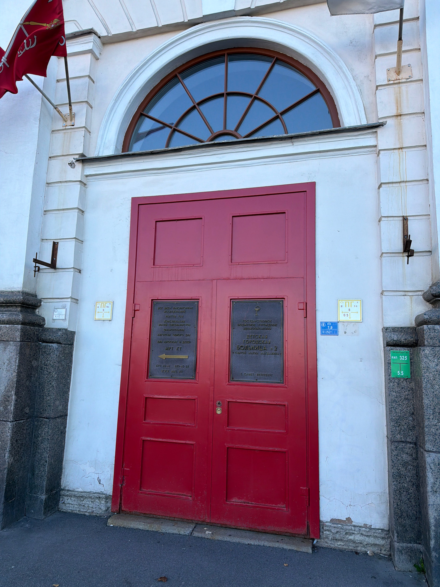 The image shows a bright red double door on a white facade building. Above the door is a semicircular window with wooden mullions. The building appears to be a historical structure, likely a public building or institution, given the signage. One sign on the door reads "ДЕТСКАЯ БОЛЬНИЦА № 2" (Children's Hospital No. 2). Another sign indicates "ВСЕМ ДНЕМ ВЫСОКОКОМПЕТЕНТНЫМ ИССЛЕДОВАТЕЛЬСКО РЕНТГЕН-ТЕРАПЕВТИЧЕСКИМ КОНСУЛЬТАЦИЯ..." (All-day highly competent research X-ray therapeutic consultation...). The hospital is located in Saint Petersburg, Russia, as indicated by the text "С. ПЕТЕРБУРГЪ" (Saint Petersburg) on the signage. There is a red flag with a symbol on a flagpole to the left of the door. Two stone columns are visible on either side of the entrance. The ground in front of the building is paved.