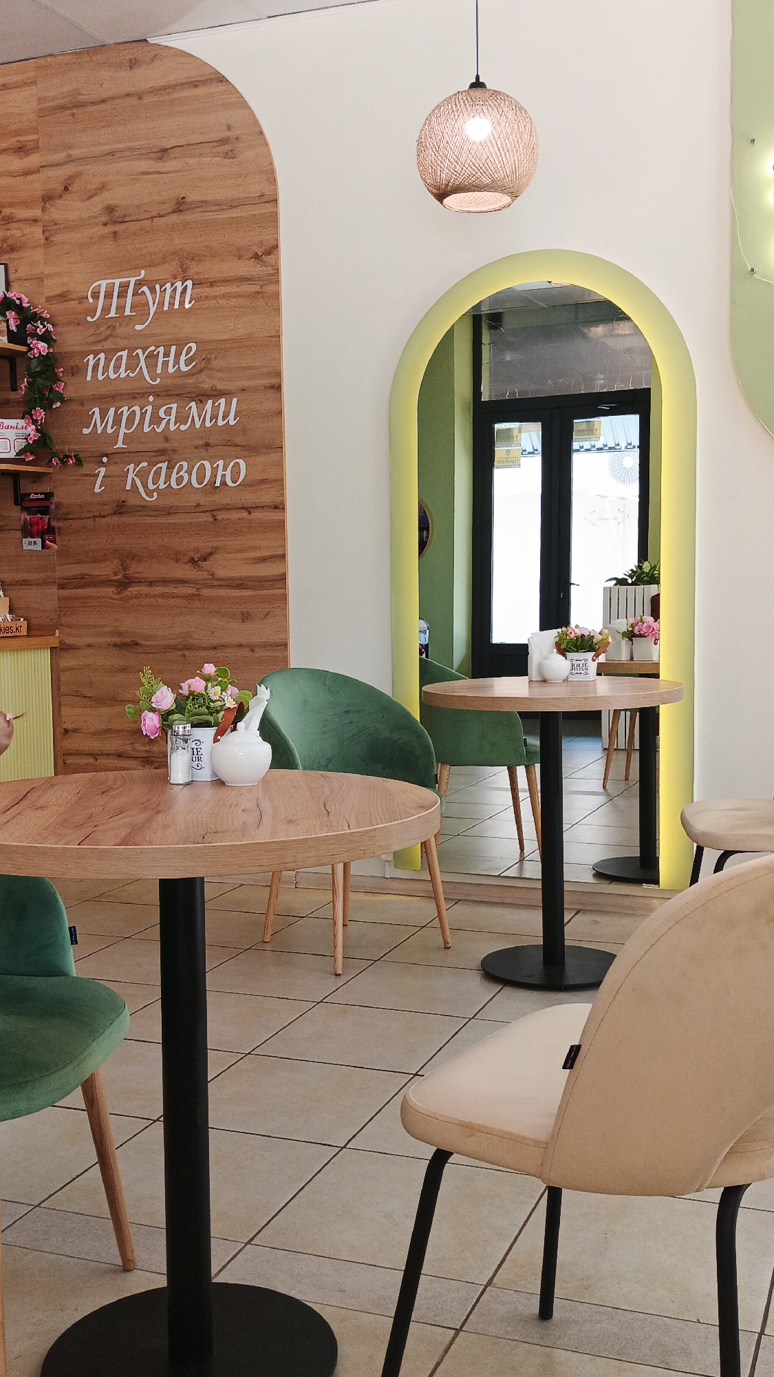 This is a medium shot of a cafe interior, featuring tables and chairs. The scene is well-lit, suggesting daytime. The decor is modern with a rustic touch, including a wooden accent wall with stylized lettering.
The main subjects are furniture: several round wooden tables with black bases and a variety of chairs. Some chairs are upholstered in plush green velvet, while others are lighter in color with black metal legs. A woven pendant light hangs from the ceiling, adding a cozy element.
Notable details include a decorative floral arrangement on one of the tables, featuring pink and white artificial flowers. A white ceramic pitcher and salt and pepper shakers are also visible on the same table. Through an arched doorway in the background, another area of the cafe is visible, with more tables and chairs and a glimpse of an exit with doors.
The overall atmosphere appears calm and inviting. The text visible on the wooden wall is in Ukrainian and reads "Тут пахне мріями і кавою," which translates to "It smells like dreams and coffee here." There are also some small labels on a shelf to the left of the wooden wall. The flooring is tiled. The location context is Kryvyi Rih, Ukraine, which is a city in central Ukraine.