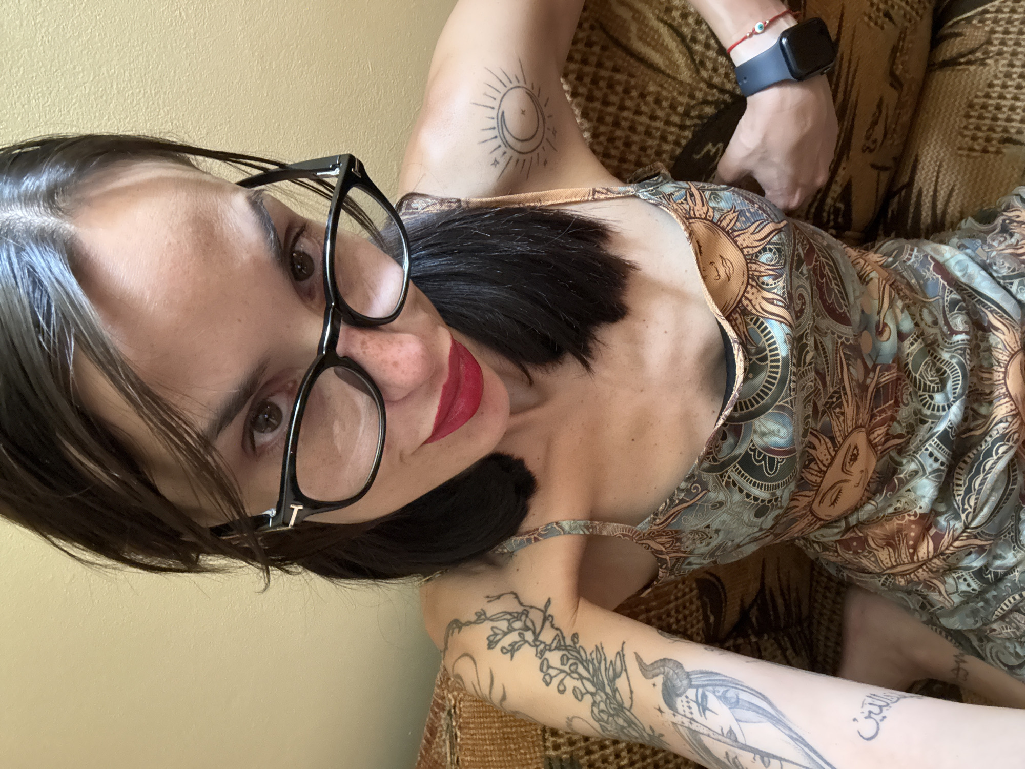 A woman with dark hair and glasses, wearing a patterned dress and makeup, is pictured from the chest up. She has several tattoos on her arms, including a bird and a sun with a face. A smartwatch is visible on her left wrist, and a red bracelet with an evil eye charm is on her right wrist. The background consists of a textured surface, likely a couch, and a plain wall. The image appears to be a selfie taken indoors. There is no information available to determine the location in the city of Pleven, Bulgaria.