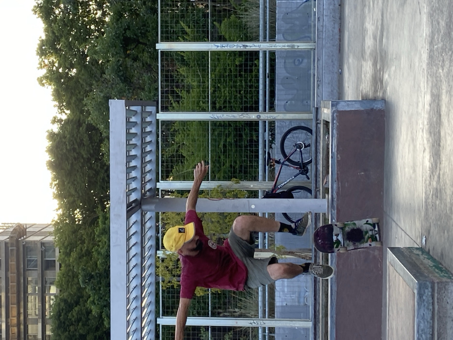 A skater in a yellow hat and maroon shirt attempts a trick at a skatepark. A bike leans against a metal grid. The skater, in mid-air, has one arm extended upwards. Trees and buildings are in the background.